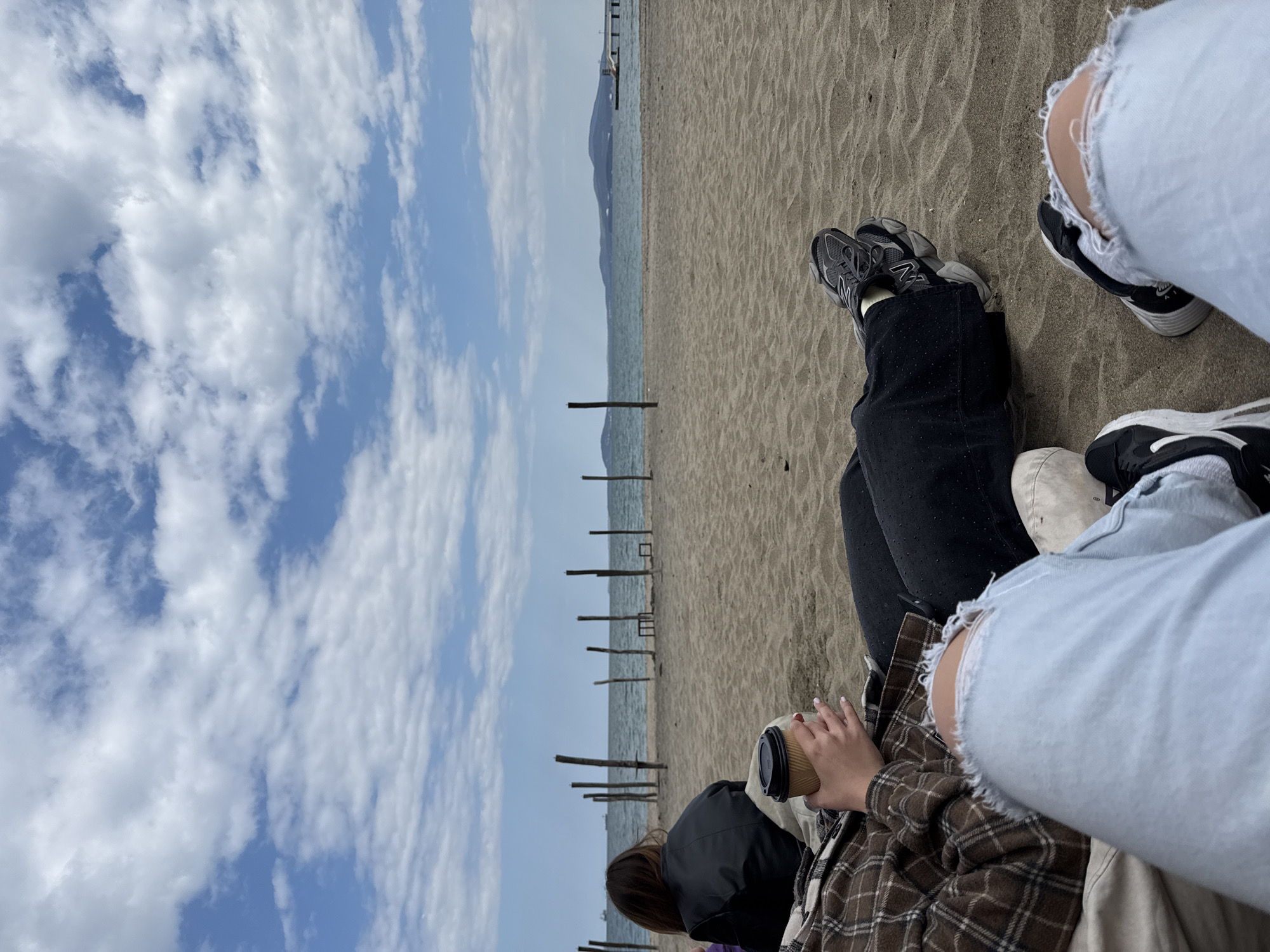 A photo taken on a beach in Burgas, Bulgaria, captures a serene yet social scene. The sky is filled with white clouds. In the distance, a pier extends into the sea. Two people are visible. One is wearing black sneakers and dark jeans, holding a coffee cup. The other wears distressed jeans and black and white sneakers. The background features the sea and a line of wooden posts.