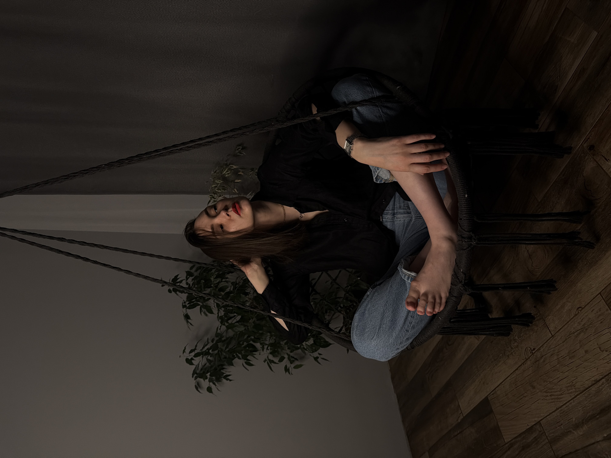 A woman is sitting in a suspended chair within an indoor space. She has brown hair and is wearing a black shirt and jeans. The chair is suspended by ropes, and a potted plant is visible behind her. The background consists of a wall and a wooden floor. The photo was presumably taken in a city. The exact address in Chaikovsky, Russia, is unknown.
