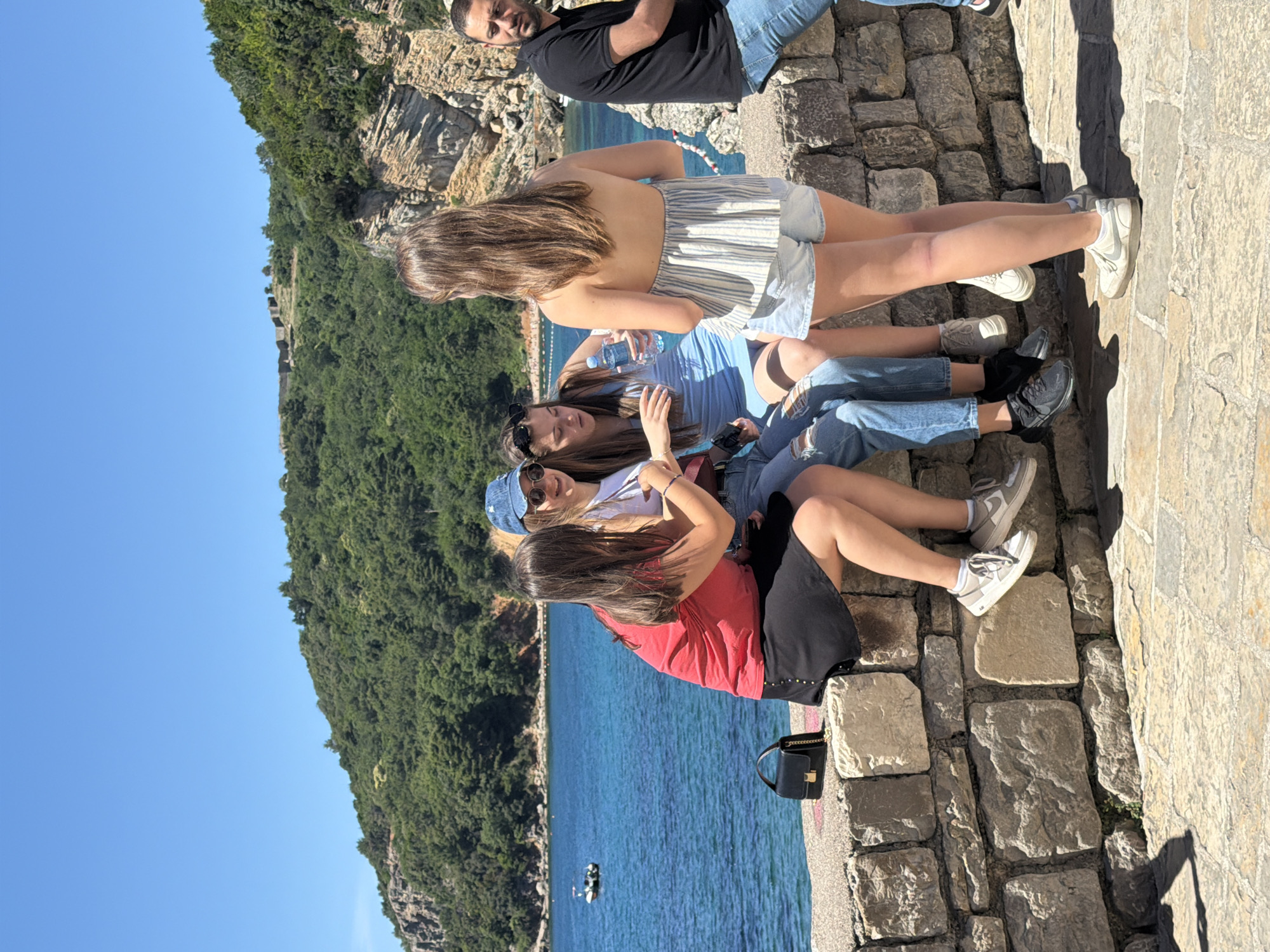 Four women and a man are sitting on a stone wall overlooking the Adriatic Sea near the Mogren Fortress in Budva, Montenegro. The women are positioned in the foreground, with varying casual summer clothing. One woman holds a water bottle. The man is in the background, wearing a black t-shirt and jeans, his face partially visible. The background includes dense green vegetation along the coastline and clear blue sky. A small boat is visible on the water. A black purse rests on the wall near the women.
