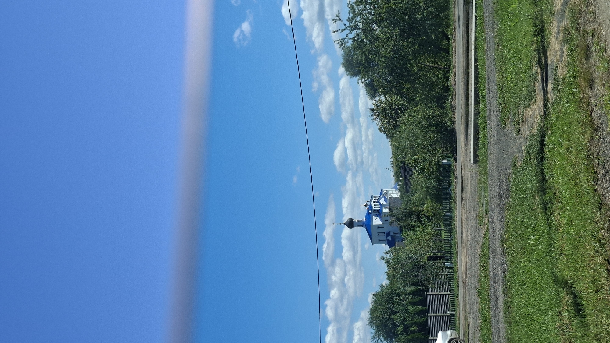 An eye-level, full shot captures a bright, sunny day with a clear blue sky dotted with fluffy white clouds. A large building, appearing to be a church, stands prominently in the center of the frame, its white walls contrasting with bright blue roofs and domes, one topped with a cross. The church is nestled amongst lush green trees, some of which are tall and full, others smaller and bush-like. In the foreground, a dirt road or pathway is visible, with patches of grass growing on either side. A section of a wooden fence and a white car can be seen on the left side of the image. A power line stretches horizontally across the upper portion of the frame, obscuring a small part of the sky. The overall scene suggests a peaceful rural or suburban setting in the city of Myadzyel, Belarus.