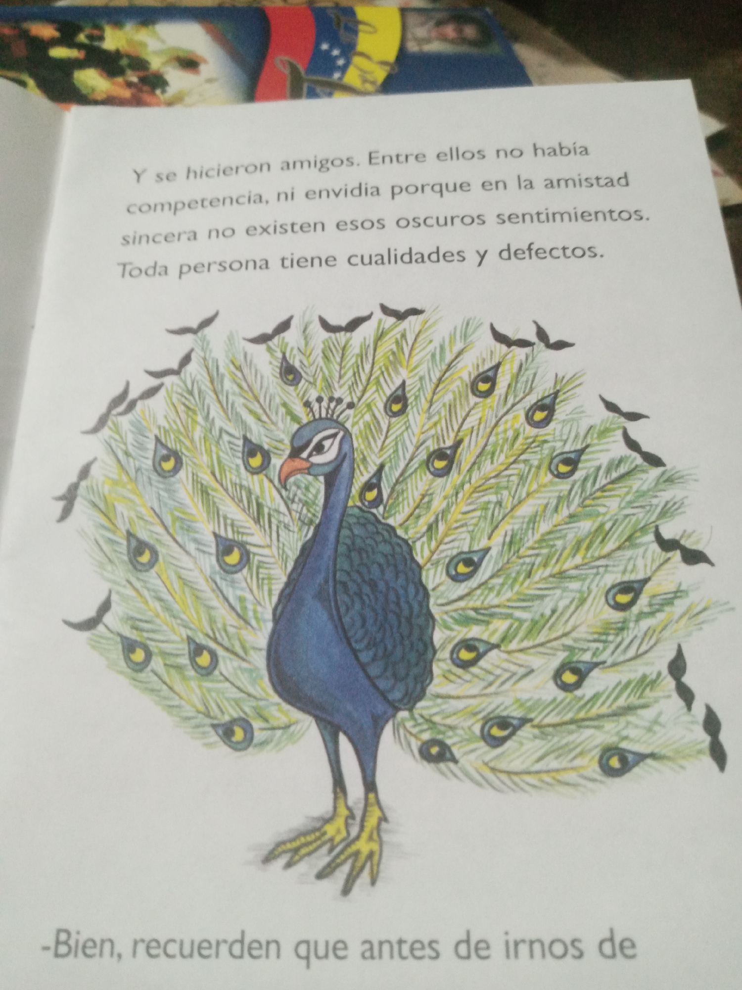 The image shows an open book with text and an illustration of a peacock. The text, in Spanish, discusses friendship, competence, envy, and personal qualities. Below the illustration, the text continues, also in Spanish, with a phrase that translates to "Well, remember that before we leave."
The illustration depicts a vibrantly colored peacock with its tail feathers fanned out. The peacock is predominantly blue and dark green, with yellow accents on its feet and crest. The tail feathers are detailed with numerous eye-like patterns in shades of blue, yellow, and black, set against a background of green and yellow lines representing the feathers. Scattered around the peacock's display are small black shapes that appear to be birds in flight.
The book itself is positioned on a surface that shows part of a colorful, patterned fabric, possibly a tablecloth or a blanket. In the upper background, a portion of a flag with red, yellow, and blue stripes and stars is visible, which resembles the Venezuelan flag. The presence of the Venezuelan flag suggests a possible location within Venezuela. However, there is no specific information within the image that can pinpoint the exact location in the city of Santa Ana, Venezuela. The image is a close-up view, and the environment outside of the book and the flag is not detailed enough to provide further context. There are no people visible in the image.