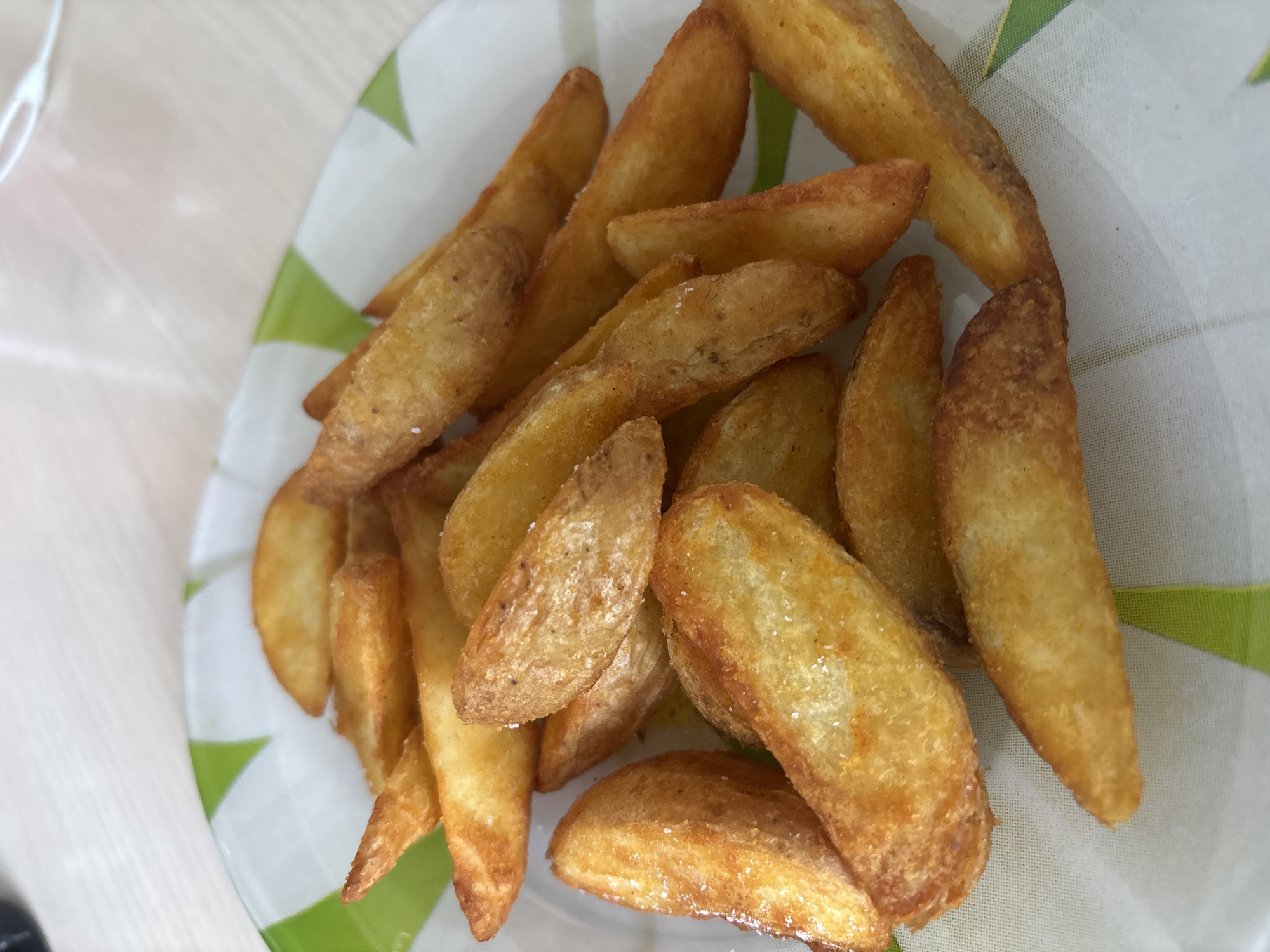A close-up, overhead view depicts a serving of freshly prepared potato wedges arranged on a plate, located in the city of Rusanovo, Russia. The potato wedges are golden-brown with varying shades of crispness, showing evidence of light seasoning, possibly salt, visible as small white crystals on their surfaces. The wedges are irregularly shaped, consistent with cut potatoes, and are loosely piled in the center of a white, possibly ceramic, plate. The rim of the plate features a distinct green and white geometric or checkerboard pattern. The background is intentionally out of focus, indicating a shallow depth of field, and appears to be a light-colored, subtly textured surface, likely a table or countertop. A blurry, indistinct light-colored object is partially visible in the upper right quadrant of the background. No individuals, specific actions, or direct interactions are discernible within the frame. The scene is illuminated by bright, even lighting, highlighting the texture and color of the food.
