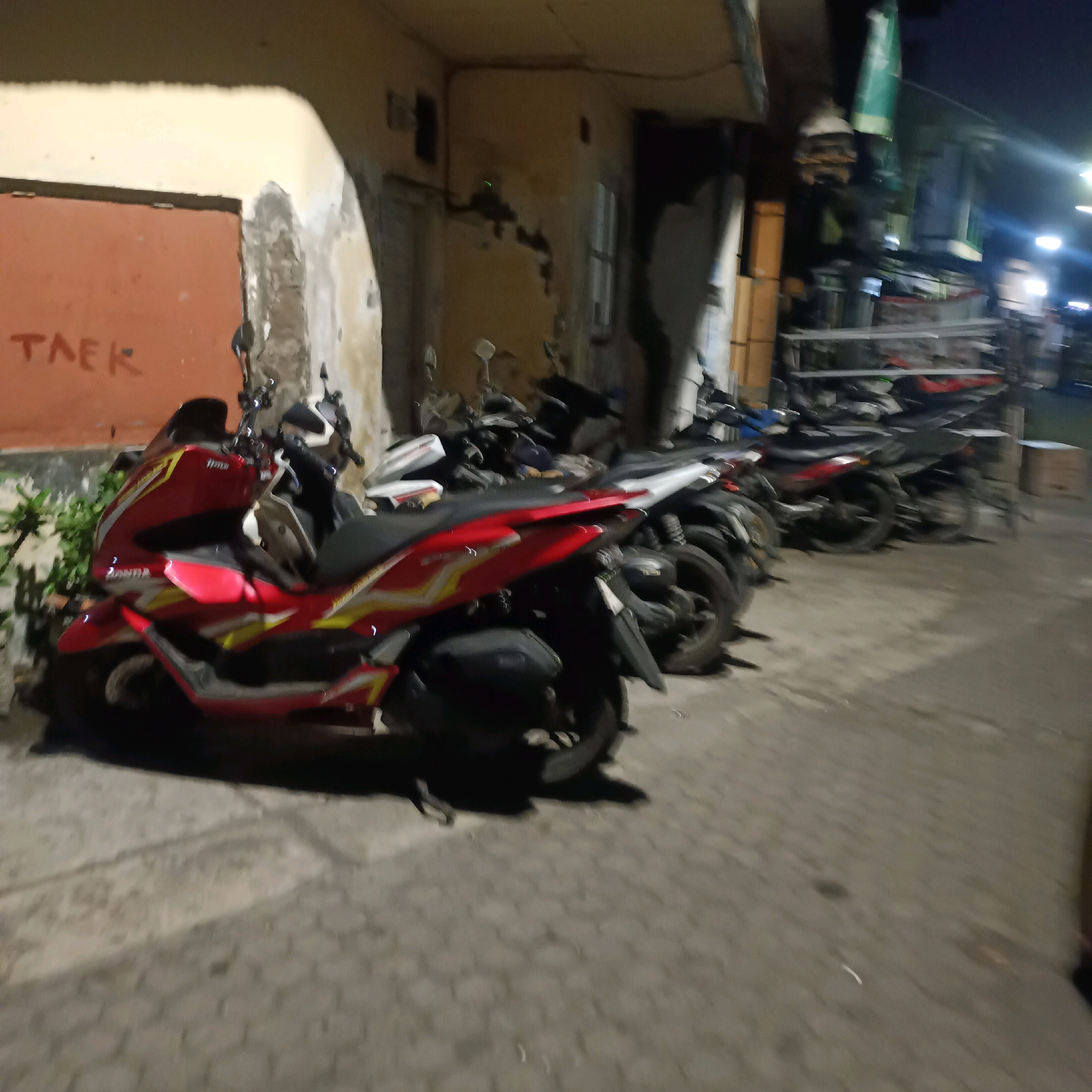 The image shows a row of parked motorcycles in what appears to be an alleyway or parking area in Surabaya, Indonesia. A red and yellow scooter is prominently featured in the foreground, parked next to other motorcycles. The background includes a building with peeling paint and a sign that reads "TAEK". The lighting suggests it is evening or nighttime, with streetlights visible in the distance. The ground is paved with cobblestones.