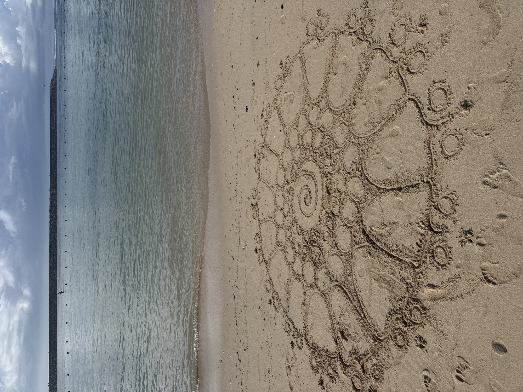 The image depicts a beach scene in Kuta Selatan, Indonesia. On the right side of the frame is the sand, where a large, intricate spiral mandala drawing has been created. The mandala consists of circles and swirling patterns, with some smudging and disruption, including footprints. The left side of the image shows the ocean with a breakwater stretching into the distance. The sky above the breakwater is visible, with some clouds. No people are clearly visible in the image, although the footprints suggest human presence.