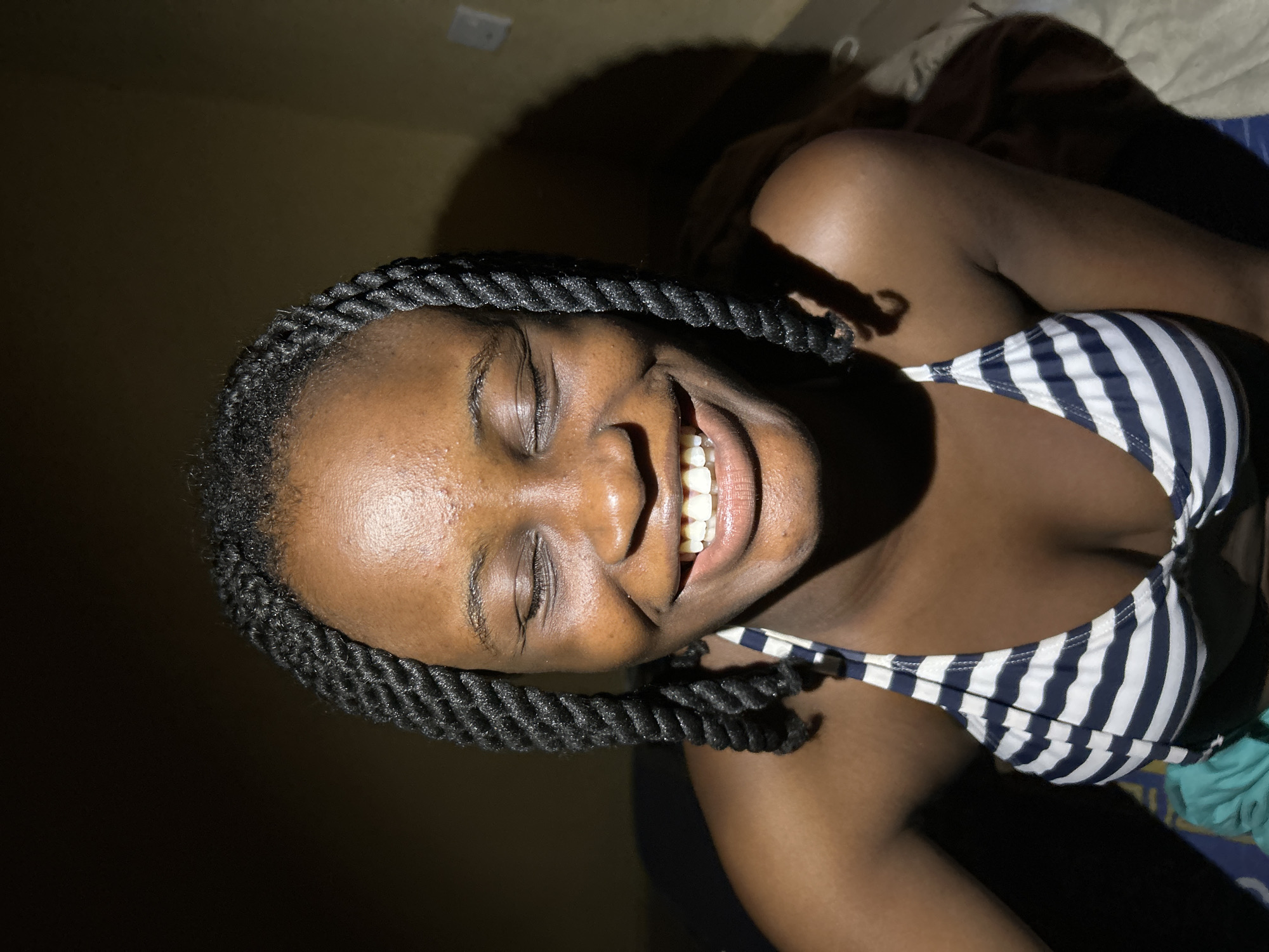 A close-up, medium shot captures a young Black woman from the chest up. She is positioned centrally in the frame with her eyes closed and a wide, genuine smile revealing her teeth. Her dark, braided hair frames her face. She is wearing a blue and white striped bikini top. The background is dark and out of focus, with a hint of a yellow wall on the right side, suggesting an indoor setting. The lighting is dramatic, casting shadows on her face and body. The image appears to be taken in Ihiagwa, Nigeria.