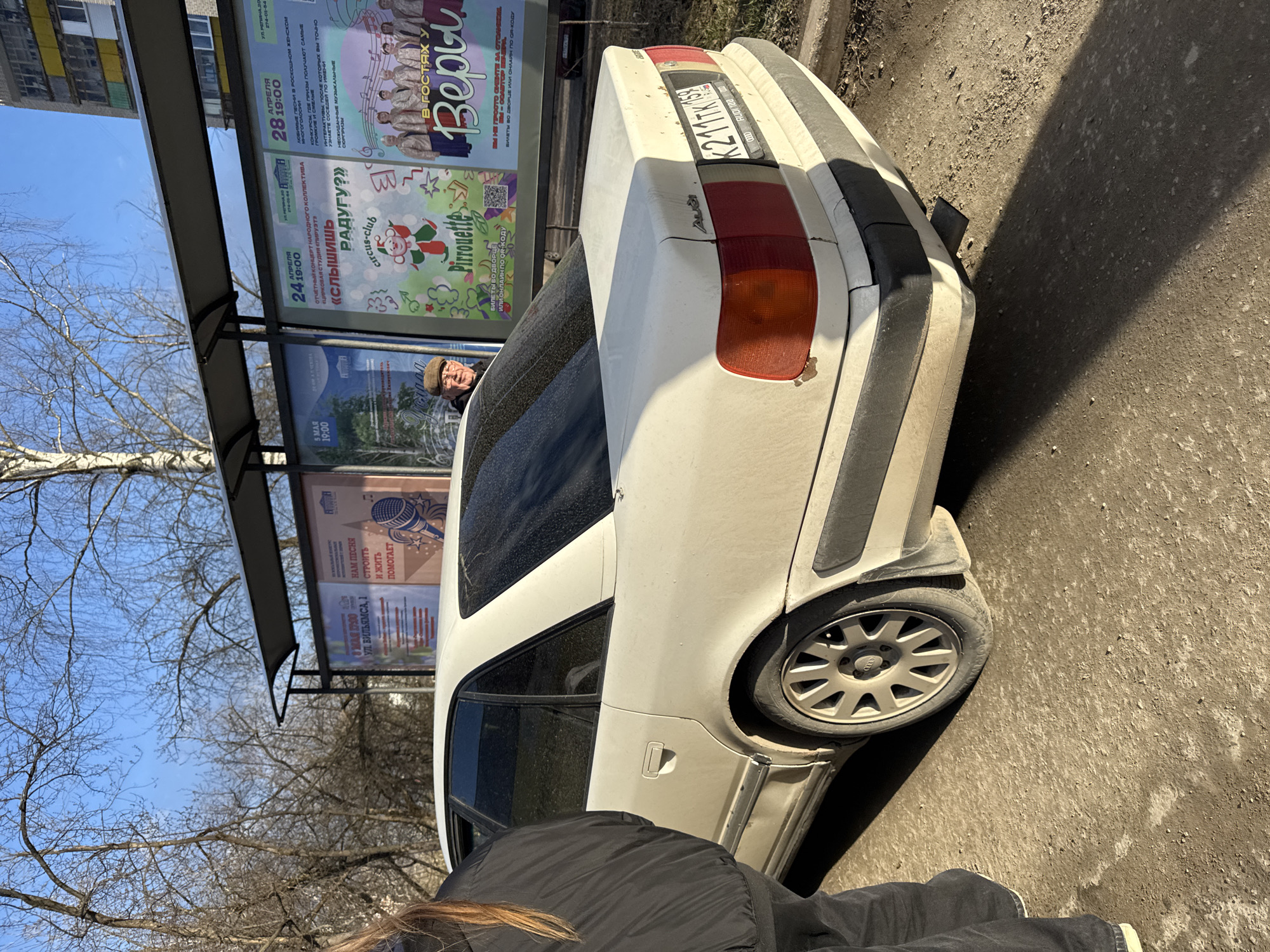 This outdoor, upright image captures a street scene in what appears to be Perm, Russia, given the license plate and text. In the foreground, the back of a person's head and body, dressed in a black jacket, is visible. The central subject is a white, somewhat dirty Audi sedan, seen from its rear-left side, parked on an asphalt surface next to a curb. The car's Russian license plate reads "K211TK 159", with "159" being the region code for Perm Krai. An Audi logo is also visible on one of the car's wheels.
Inside the car, through the rear window, a man wearing a cap is seen looking out. The car shows signs of wear, including rust near the taillight. To the left of the car is a bus stop or shelter structure, featuring multiple advertising posters in Cyrillic.
The bright sunlight casts strong shadows, suggesting it is daytime, possibly late morning or afternoon. The bare trees indicate late winter or early spring weather. The sky is clear and blue.
Visible text on the posters advertises various events:
- A poster for "В гостях у Веры" (Visiting Vera) mentions prizes and surprises.
- Another poster advertises "Цирковая студия «Пируэт»" (Circus Studio "Pirouette") and asks "Слышишь РАДУГУ?" (Do you hear the RAINBOW?).
- A poster mentions an "Отчётный концерт народного коллектива" (Reporting Concert of the National Collective).
- Event dates and times include "24 АПРЕЛЯ 19:00" (April 24, 19:00), "28 АПРЕЛЯ 19:00" (April 28, 19:00), and "5 МАЯ 19:00" (May 5, 19:00).
- Addresses "УЛ. РЕТИНА 20" (Retina St 20) and "УЛ. ВИЛЬЯМСА, 1" (Williams St 1) are present, along with phone numbers "274-05-04" and "274-01-64".
- A slogan "НАМ ПЕСНЯ СТРОИТЬ И ЖИТЬ ПОМОГАЕТ" (Song helps us build and live) is also visible.