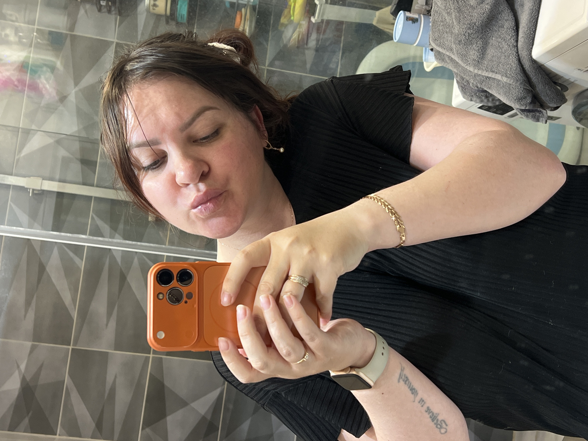 A woman is taking a selfie in a bathroom. The woman has brown hair pulled back with a clip, and she is wearing a black top. She is holding an orange smartphone in her hands, and she has rings on her fingers. She is wearing a gold bracelet on her wrist and a smartwatch. The background shows gray geometric-patterned tiles on the wall, with a glimpse of bathroom fixtures and a washing machine. The location of Noviy, Russia, cannot be determined from the image.