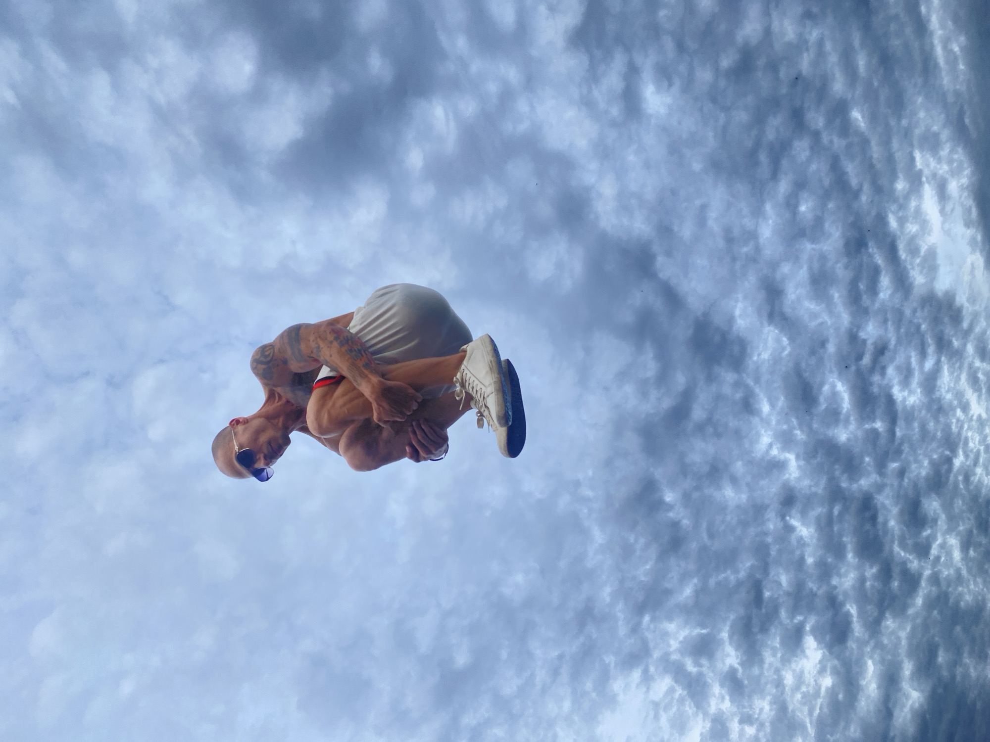 A bald man wearing sunglasses and white shorts is captured mid-air, in a crouched position, against a backdrop of a cloudy sky. The man has tattoos on his arms and is wearing white sneakers. The perspective suggests he is jumping upwards. The image is taken from a low angle looking up at the man. The sky is filled with textured, greyish-blue clouds, suggesting an overcast or stormy day. There are no discernible city landmarks or specific architectural features visible that would indicate a location within Pleven, Bulgaria.
