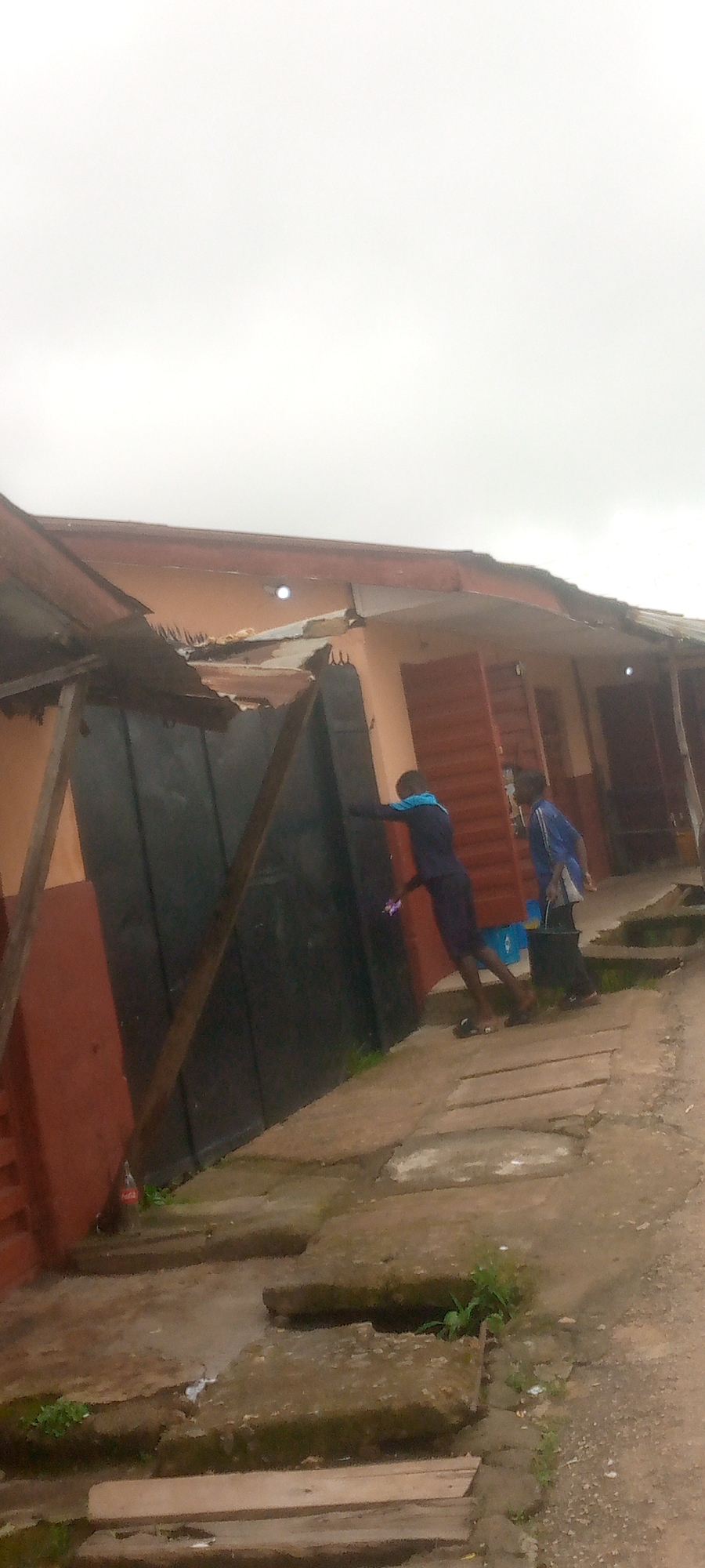 In an outdoor urban scene, two young individuals are positioned near a dark, corrugated metal gate. The gate is attached to a building with an orange-hued facade and a damaged, peeling roof. The sky is overcast and bright white.
One individual, wearing a dark blue long-sleeved shirt with a light blue collar and dark shorts, leans against the gate, their right hand resting on it. They appear to be holding a small, light-colored object in their left hand.
The second individual, standing to the right of the first, wears a blue long-sleeved shirt and dark pants. They are carrying a black bucket in their right hand and a white object in their left hand. This individual is looking towards the right.
The ground is paved with uneven, rectangular concrete slabs interspersed with dirt and patches of green grass. A Coca-Cola bottle stands near the base of the building to the left of the gate. The overall scene suggests a modest urban environment, possibly a residential or commercial area with some signs of disrepair.
