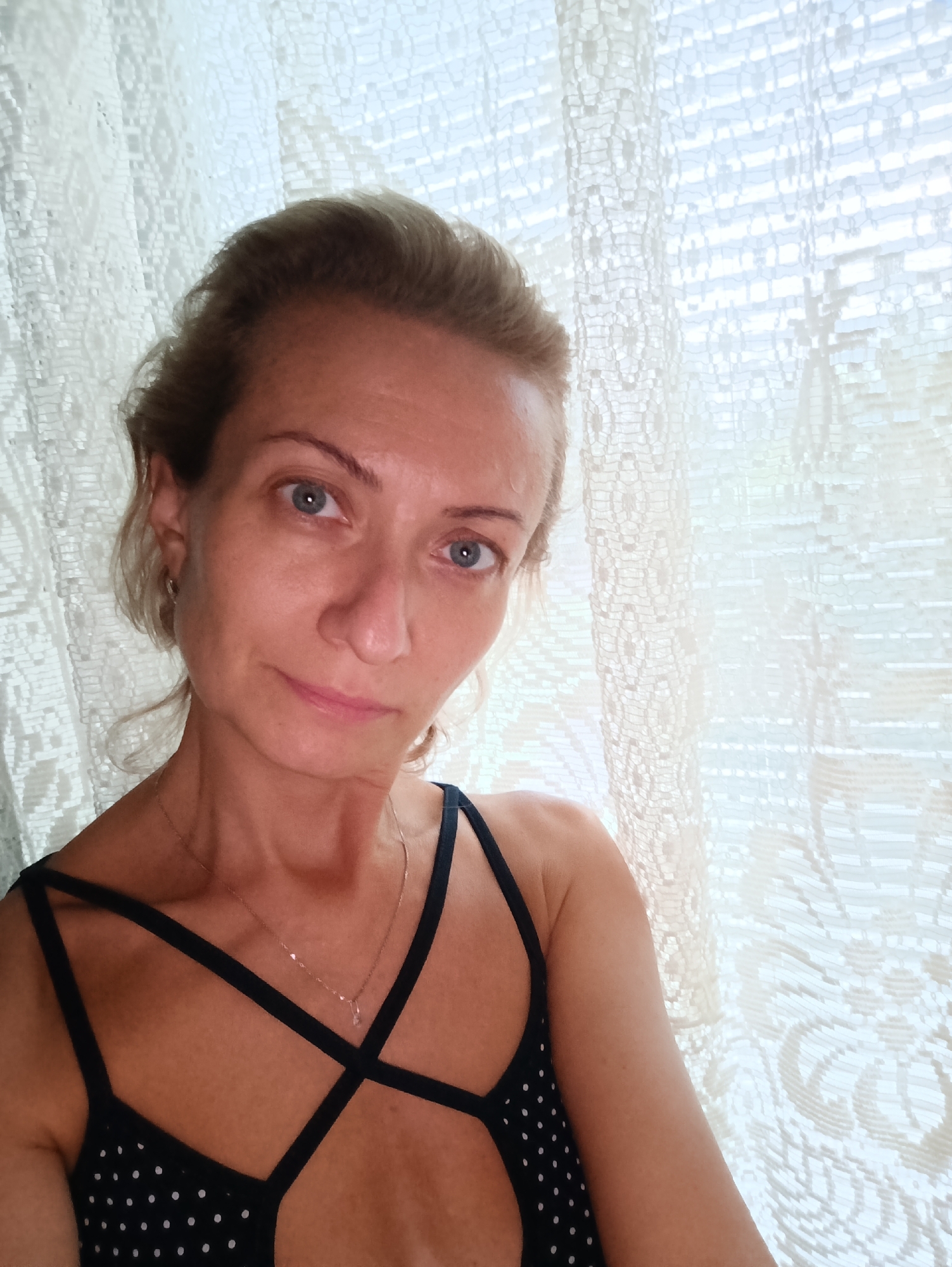 A woman with blonde hair and blue eyes is taking a selfie in front of a lace curtain. She is wearing a black polka-dot top with crisscross straps and a delicate silver necklace. Her hair is pulled back loosely, with some strands framing her face. The lighting suggests it is daytime, with natural light filtering through the curtain. The background is a white, patterned lace curtain, and there is a hint of what looks like a window or another curtain behind it. No specific location in Moscow, Russia can be identified from the image.