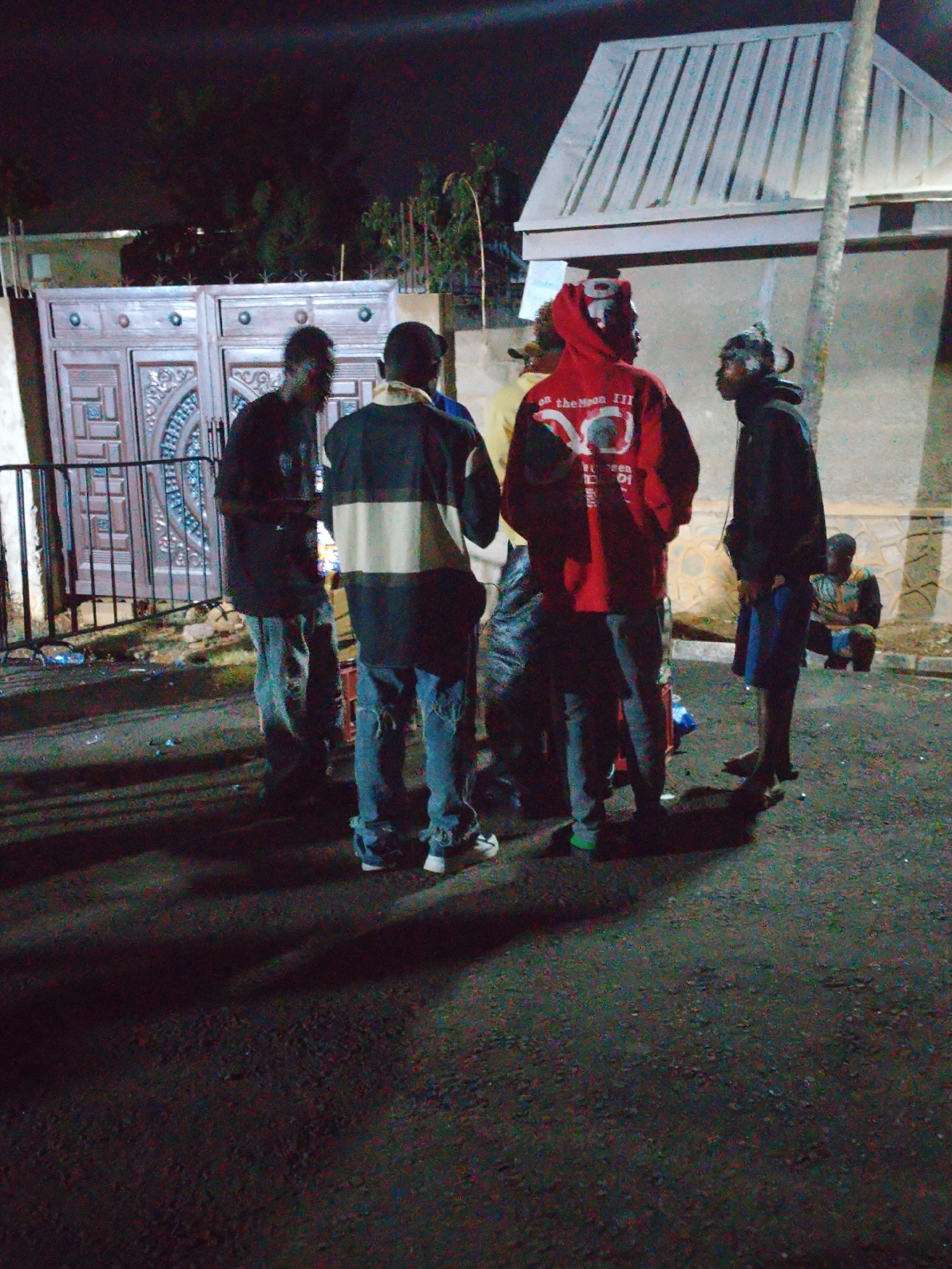 In the city of Unknown, Unknown, a group of approximately six individuals are gathered outdoors at night. The scene is dimly lit, casting long shadows on the paved surface. The group is positioned near a large, ornate wooden gate and a building with a slatted roof. Some individuals are standing, while one person is seated. The standing individuals are wearing a variety of clothing, including hoodies, jeans, and t-shirts. Their actions and interactions are not clearly discernible due to the lighting and distance. The environment appears to be urban, with buildings and landscaping visible in the background. Litter and debris are scattered on the ground.
