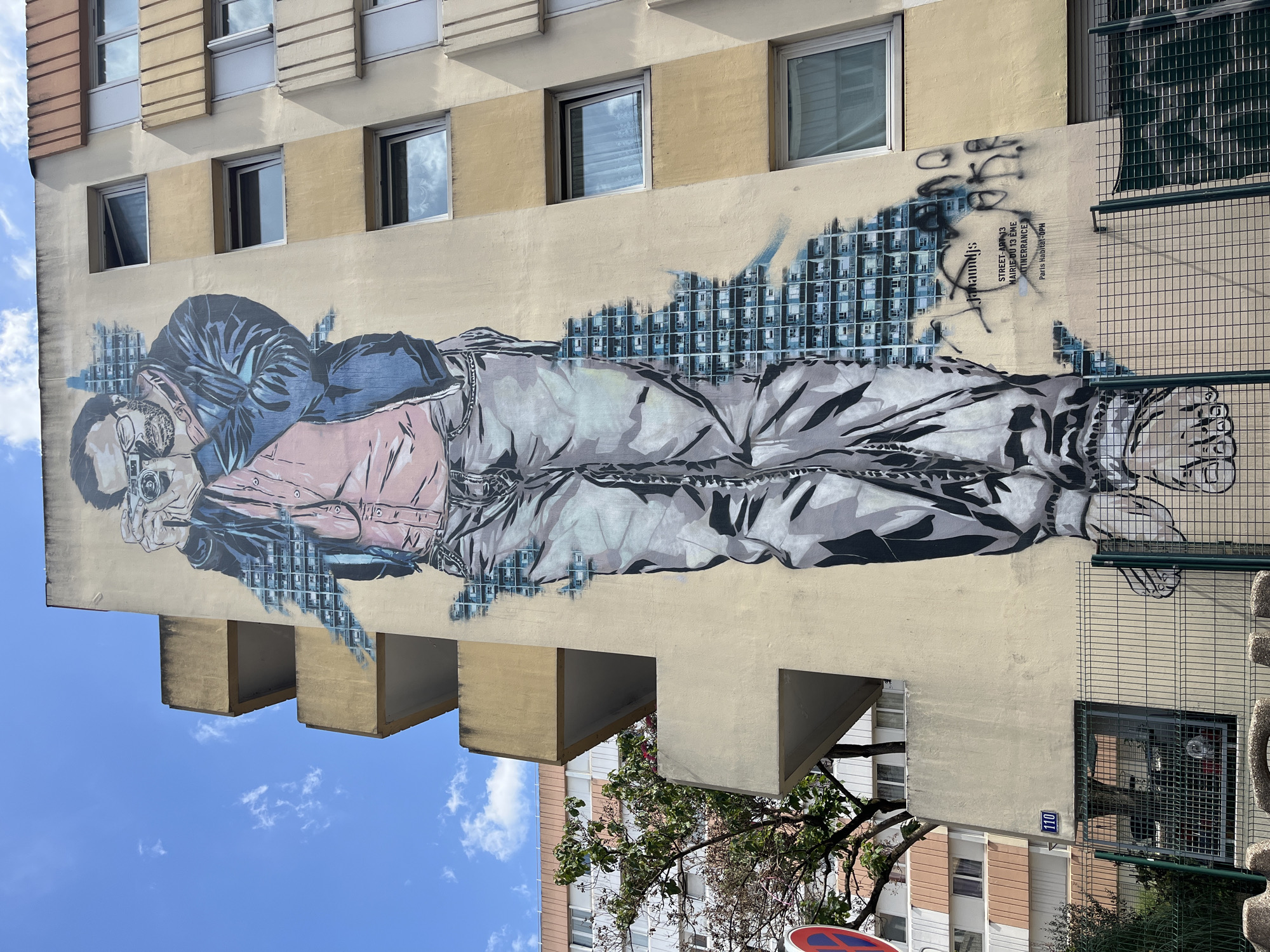 The image shows a large mural of a man holding a camera on the side of a light yellow building in Paris, France. The mural is painted in a realistic style with detailed shading and textures. The man is wearing a blue jacket, a pink shirt, and grey jeans, and he is looking through the viewfinder of his camera. The mural is positioned on the wall of an apartment building, with several windows visible to the right of the artwork. To the left of the mural, there are concrete balconies and a green tree with pink flowers. In the foreground, a metal fence runs along the bottom of the image, partially obscuring the lower portion of the mural and the base of the building. The sky above is blue with scattered white clouds. There is also a street sign with the number 110 on the building. Text on the mural identifies it as "STREET-ART 13 MAIRIE DU 13EME ITINERANCE Paris Habitat-OPH." The mural appears to be part of a street art initiative in the 13th arrondissement of Paris.