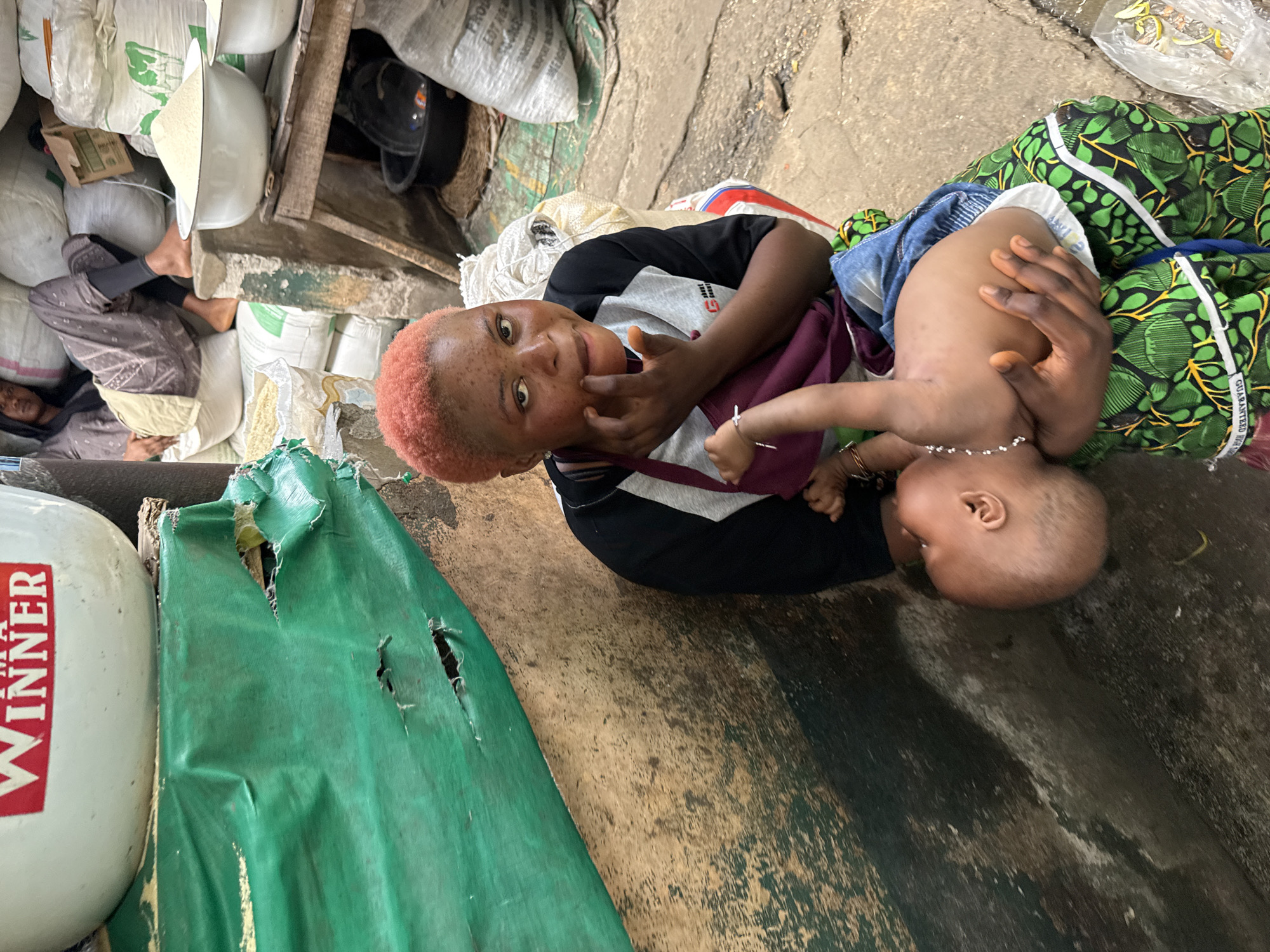 This close-up, eye-level shot captures a woman holding a baby in what appears to be a bustling market or storage area in Lagos, Nigeria. The image is oriented sideways, but depicts the subjects looking towards the viewer.
The main subject is a woman with distinctive short, reddish-orange hair, wearing a black, grey, and white t-shirt. She is smiling directly at the camera, with one finger near her mouth. She cradles a baby, possibly a toddler, dressed in denim shorts over a diaper and wearing a light-colored beaded necklace. The baby's head is visible, turned away from the camera. The woman's arm, adorned with a simple bracelet, is visible, securing the child.
The background is filled with numerous large, white sacks, likely containing grains or other commodities, stacked high. Behind the woman and baby, a vibrant green and black patterned fabric, possibly a blanket or wrapper, is visible. In the deeper background, another person is seen lying or resting amidst the sacks, suggesting a makeshift sleeping or resting spot within the crowded environment.
To the left of the frame, a white cylindrical container with the red text "WINNER" is partially visible. A torn, dark green tarp or plastic sheet with holes lies on the ground, indicating wear and tear from use. Wooden planks and uneven ground surfaces further define the rustic and functional setting. The overall scene conveys a sense of everyday life and work in an active commercial space.
