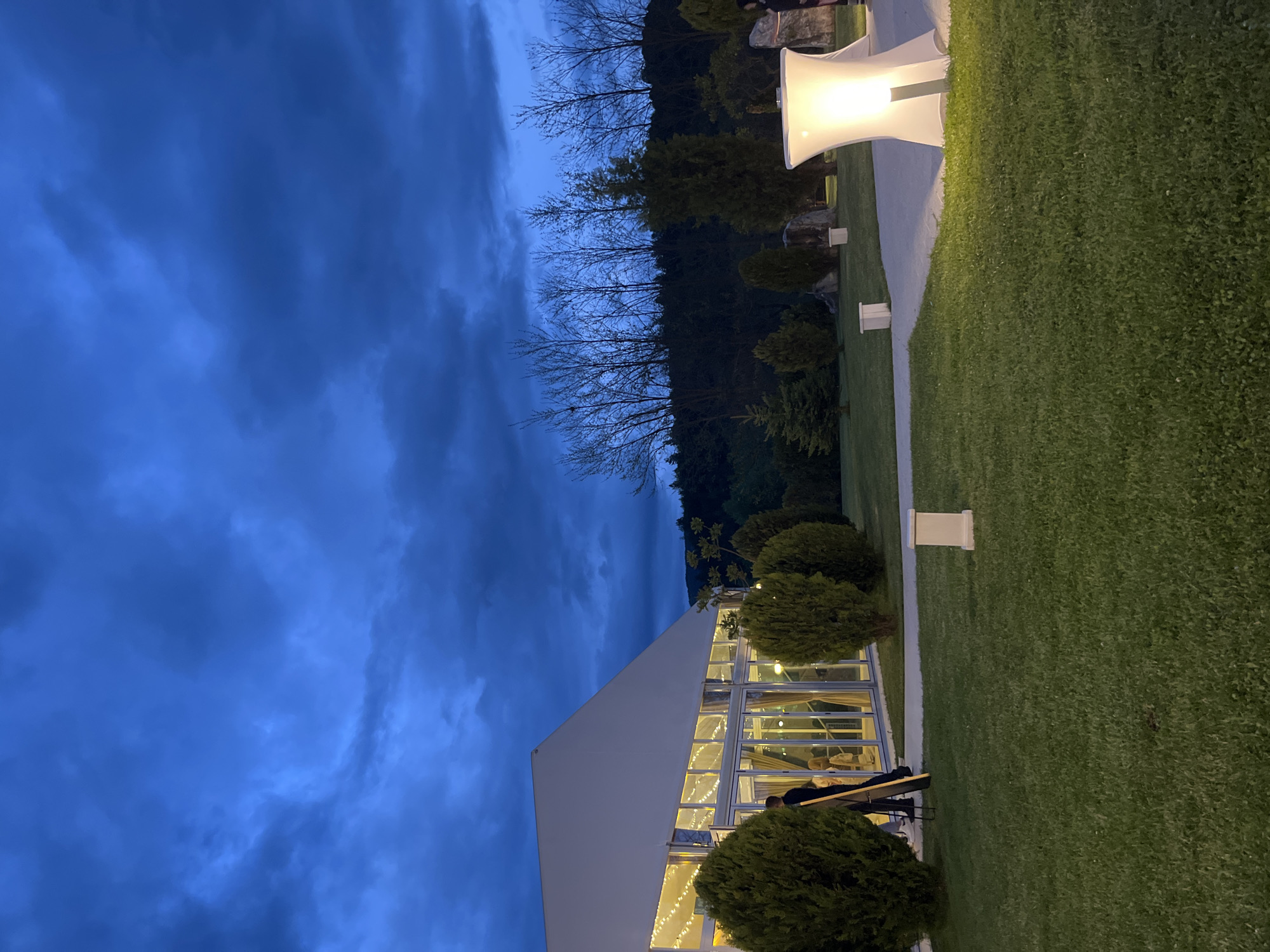 The image shows an outdoor scene at twilight, possibly in Koprivlen, Bulgaria. A large, white tent-like structure with multiple windows stands adjacent to a well-maintained green lawn. The structure is lit from within, suggesting an event. Two individuals are visible near the tent, possibly setting up or taking down equipment. White, trumpet-shaped lights are placed along a pathway. Background trees and a cloudy, dark blue sky create the atmospheric setting.