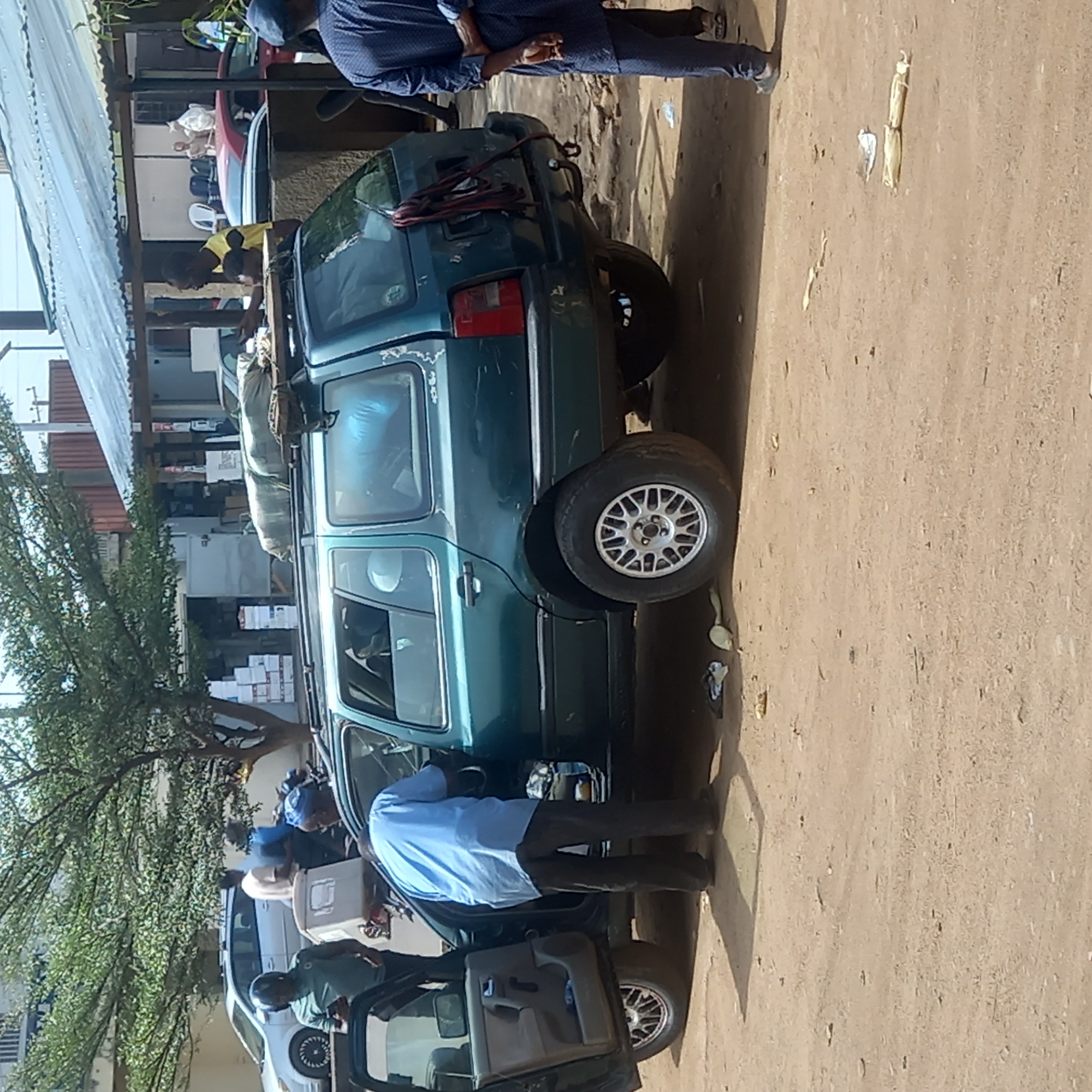 In Dadi Riba, Nigeria, a green SUV with luggage strapped to its roof is parked on a dirt lot in front of a building, possibly a commercial establishment given the boxes stacked inside. Several individuals are present. Some are dressed in light blue shirts while others are in blue-patterned shirts. One person is standing near the rear of the SUV while others are loading items into the vehicle. A tree is partially visible on the left side of the frame.