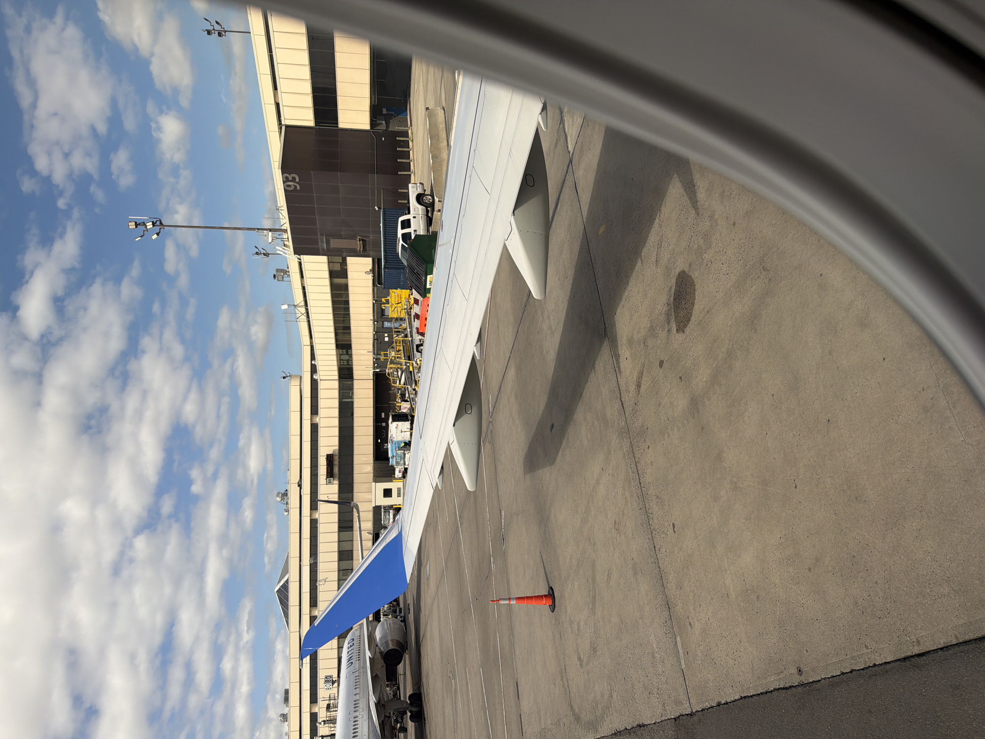 The image shows a view from inside an airplane, looking out the window at an airport scene. The wing of the airplane is visible in the foreground, with flaps extended. Beyond the wing, there is an airport terminal building with the number "93" prominently displayed. Airport ground vehicles are visible near the terminal. The sky is partly cloudy. The presence of a United Airlines plane and the gate number suggest the location is likely Newark Liberty International Airport (EWR), United States of America. An orange traffic cone sits on the tarmac in the area between the aircraft and terminal.