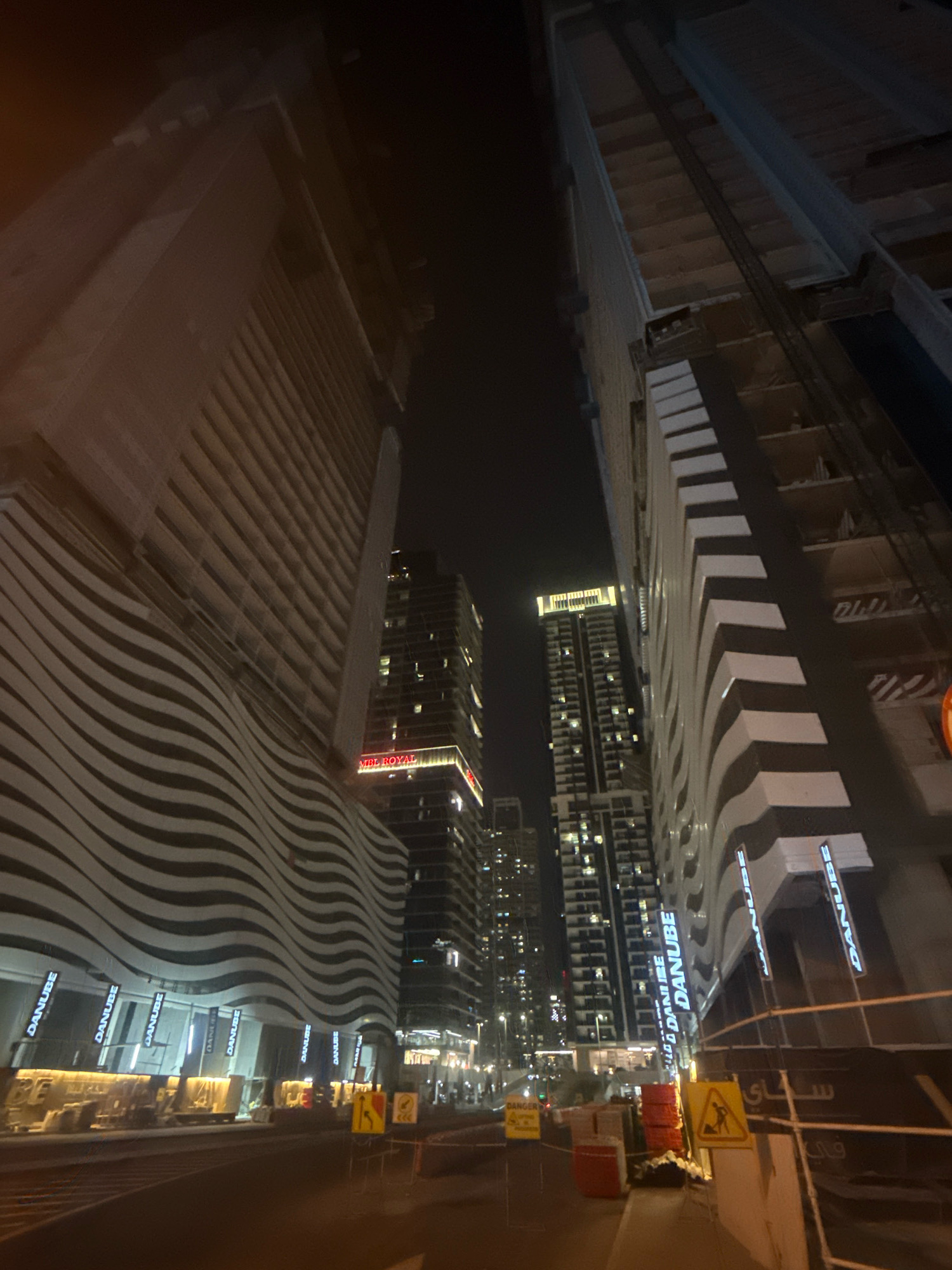This image captures a nighttime street view in the Dubai Marina district of Dubai, United Arab Emirates. The foreground features a road with traffic barriers and construction signage, including a yellow sign depicting a worker with a pickaxe. The midground is dominated by a large, modern building with a distinctive wavy facade adorned with repeating "DANUBE" logos illuminated in white. To the right of this building, another skyscraper is under construction, its framework visible. In the background, multiple high-rise buildings are lit up, indicating a densely populated urban environment. One building in the background displays a red illuminated sign that reads "VRE ROYAL". The overall scene suggests a vibrant, ongoing urban development in one of Dubai's prominent architectural hubs.