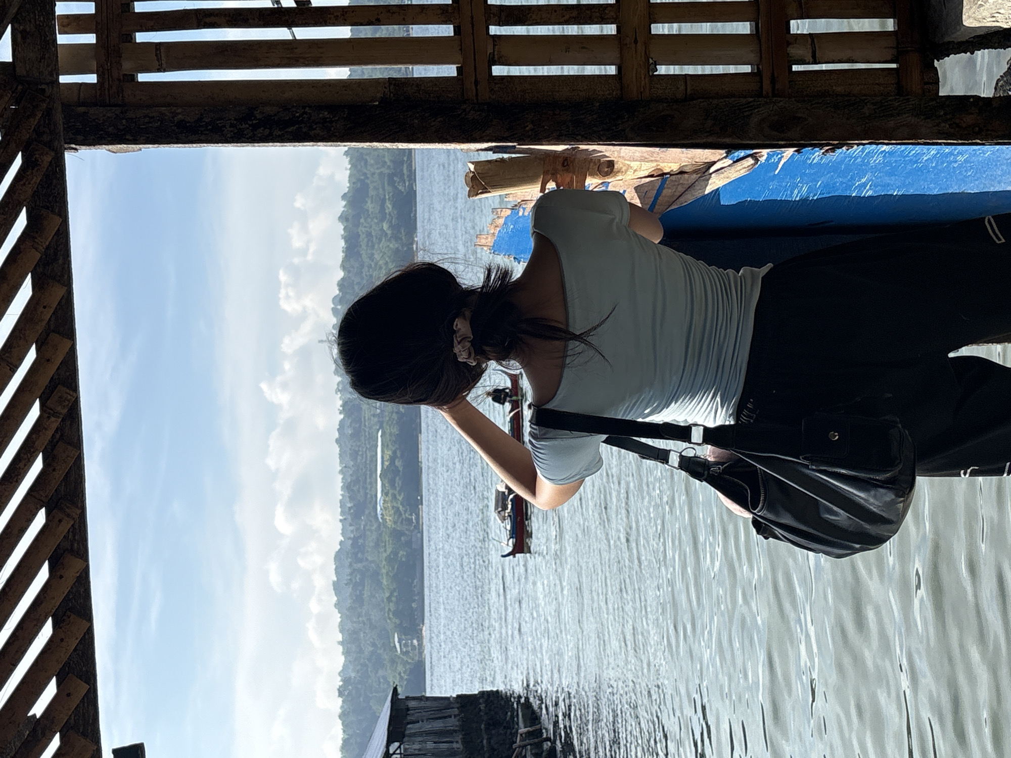 A person, facing away from the camera, stands on a wooden structure overlooking a body of water. The person has dark hair pulled back and wears a light blue off-the-shoulder top and dark pants. A black shoulder bag hangs on their left side. In the water, a small red and white boat with a person on it is visible in the mid-ground. The background features a lush, green, hilly coastline under a partly cloudy blue sky. The foreground shows wooden beams and bamboo lattice work, suggesting a rustic, open-air structure. This scene is consistent with the coastal areas of Davao City, Philippines, known for its proximity to the ocean and hilly terrain.