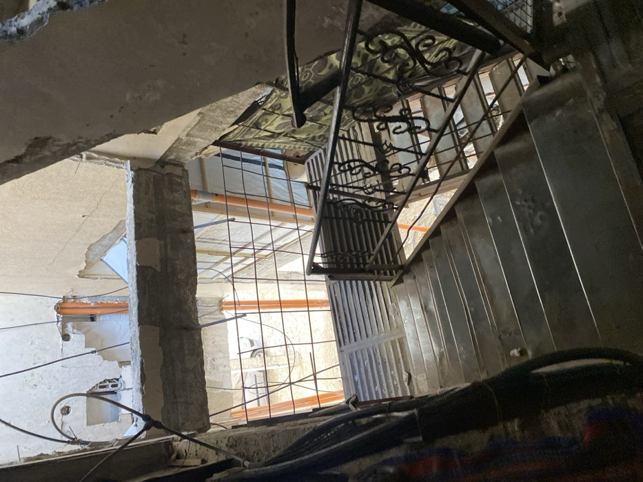 The image captures a downward view of a metal staircase within a significantly damaged multi-story building located in Khan Yunis, Palestinian Territory. The staircase features flat, worn metal treads and is bordered on the right by a black, ornate wrought-iron handrail. Overhead and across the mid-ground, substantial concrete beams are exposed, exhibiting fracturing and visible rebar, indicating structural compromise. Rough, light-colored plaster or concrete walls show extensive deterioration, with large sections missing or broken away. On the left, several black electrical cables are bundled and descend alongside the wall. Through the open lower levels, an outdoor ground area is visible, containing multiple vertical orange pipes, a dark metal grid fence, and light-colored sheeting or tarpaulin. A partial view of a white vehicle is discernible in the distant ground area. A patterned green, black, and white blanket or rug is draped over a structural element on the right side of the staircase. No individuals or specific activities are depicted.