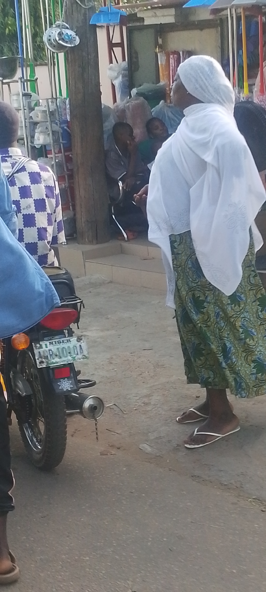 The image depicts a street scene in Lokoja, Nigeria. An elderly woman wearing a white headscarf, a white blouse, and a long green patterned skirt stands on the roadside, wearing flip-flops. To her left, there's a parked motorcycle with a license plate reading "NIGER AGR-1040A". Two other people can be seen to the left of the motorcycle, with one wearing a purple and white checkered shirt and the other wearing a blue shirt. In the background, there are a couple of individuals sitting on the steps of a building. There are also items that look like household goods displayed next to them.