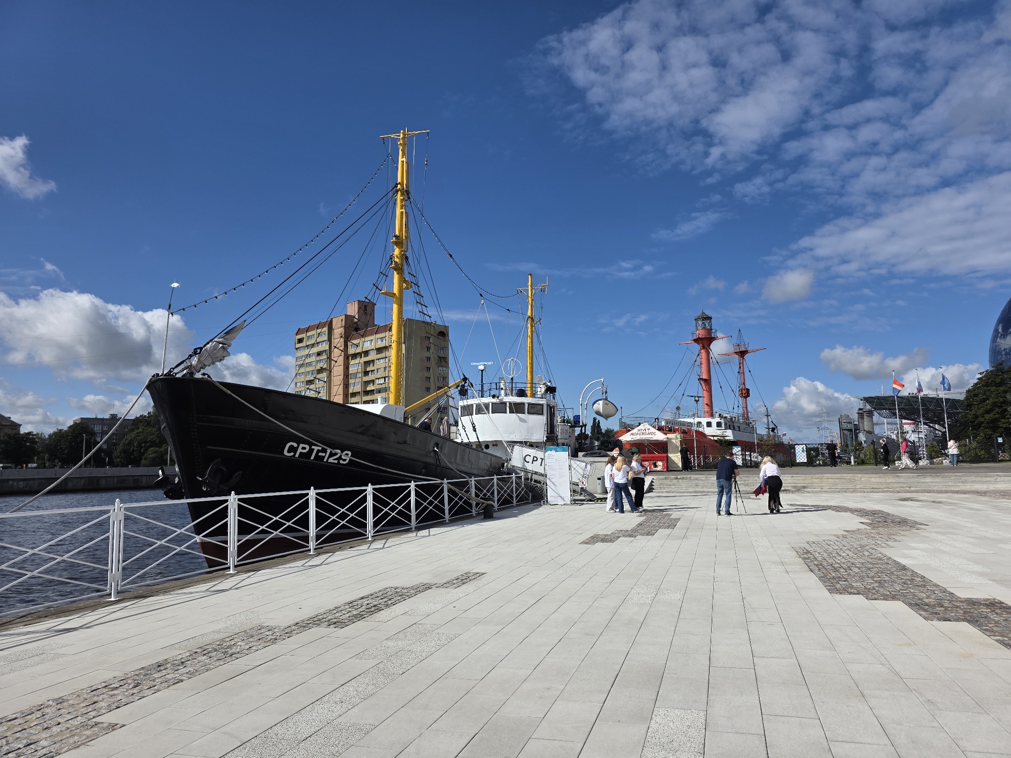 In Kaliningrad, Russia, a black ship with "CPT-129" visible on its side is docked alongside a paved waterfront promenade. The ship features a tall yellow mast with lights strung along it, and a white superstructure. Further down the promenade, a red lighthouse with a lantern room stands. Several people are walking and standing on the promenade. In the background, there are buildings, trees, and a large, futuristic-looking structure with a curved facade, possibly an aquarium or exhibition center. The sky is blue with scattered white clouds. The scene appears to be a tourist attraction or a public space in a port city.