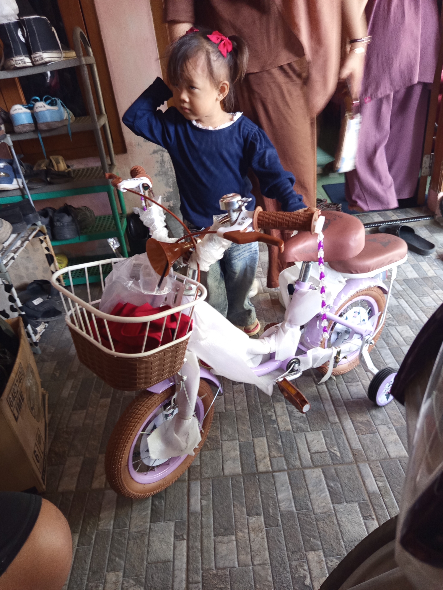A young girl with two red hair ties stands next to a lavender bicycle on a tiled floor. The bicycle has a brown wicker basket on the front containing red fabric. The girl is wearing a navy blue long-sleeved shirt and blue jeans. She is looking to her right and has her left hand on her head. In the background, two people are partially visible, wearing brown and purple clothing. A shoe rack with shoes is on the left side of the image. The scene appears to be an indoor or semi-outdoor area.