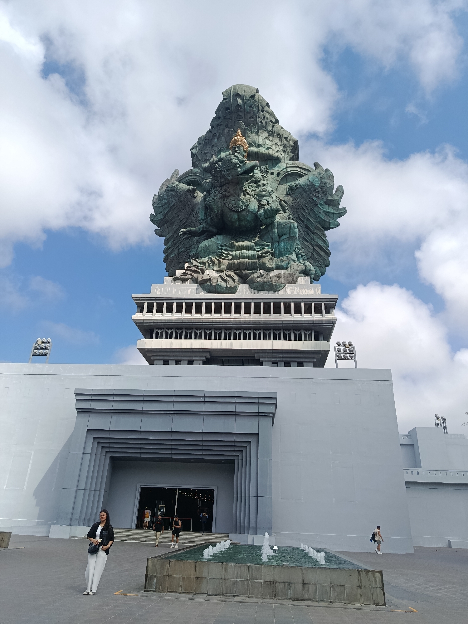 In Kuta Selatan, Indonesia, a monumental Garuda Wisnu Kencana statue looms large against a backdrop of scattered clouds and a bright blue sky. The statue, primarily a dark green, is intricately detailed, featuring wings and a golden headdress. It is mounted atop a multi-tiered, grey concrete structure with a modern architectural design, characterized by rectangular window-like openings and clean lines. In the foreground, a woman in white pants and a black jacket stands on a paved plaza, holding a small bouquet of flowers. To her right, a decorative fountain with multiple spouts adds a touch of movement to the scene. Other individuals are visible near the entrance of the building, which features a grand, recessed entryway. The overall scene depicts a tourist attraction with a focus on the impressive statue and its surrounding facilities.