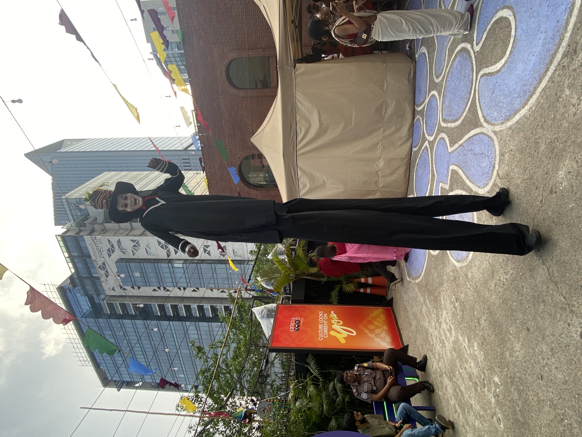 The scene depicts an outdoor event in Dhaka, Bangladesh. In the foreground, a stilt walker dressed in a black suit and a mask is visible. He stands near an advertising board with the text "Culture Looks Current on You". The ground features a painted design in blue and gray. In the background, there is a mix of people, a tent, and a modern high-rise building. Bunting is strung overhead. A security guard sits on a bench near the advertisement. The overall impression is of an event or festival occurring in an urban setting.