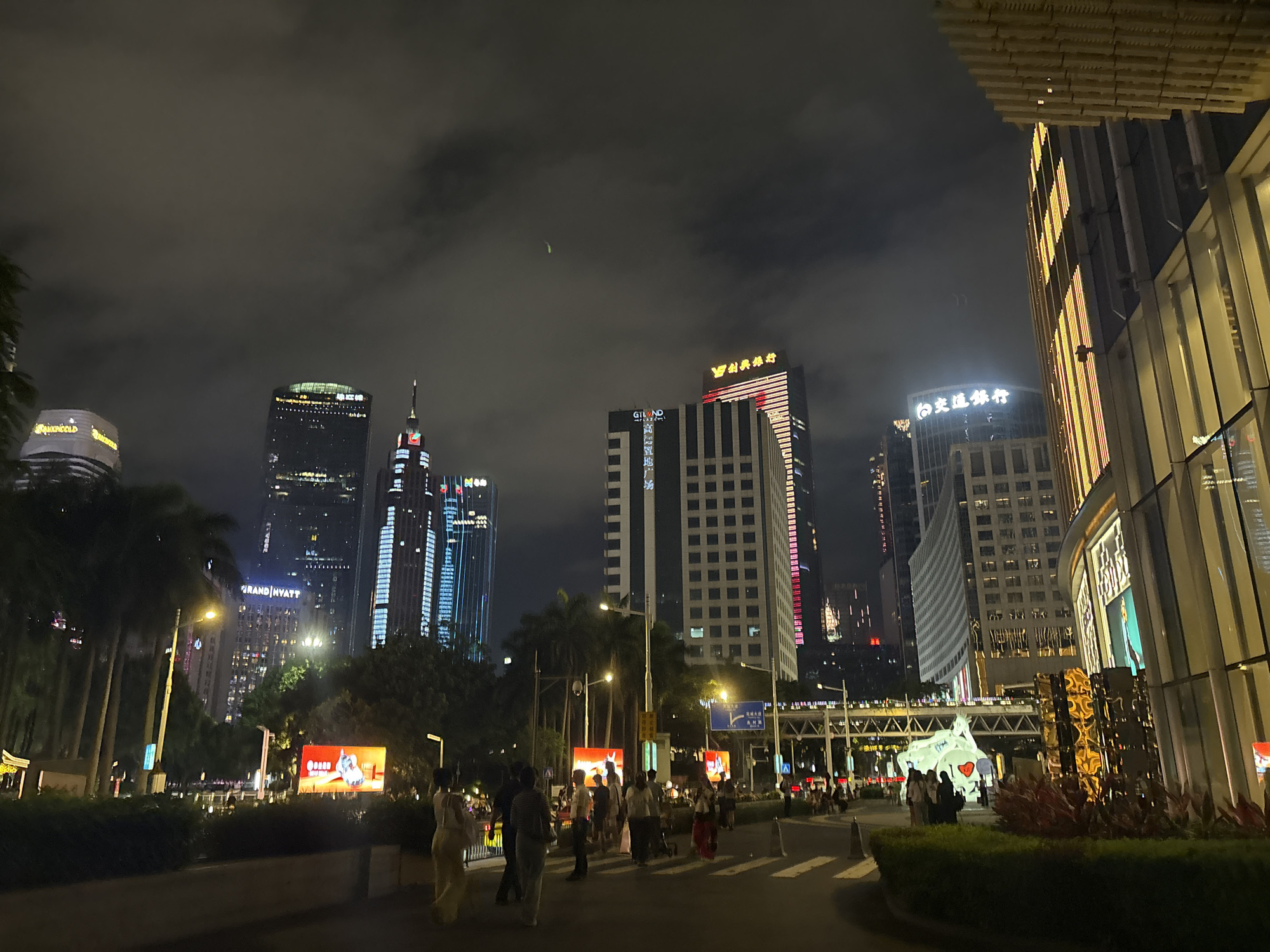 In Tianhe District, China, a nighttime scene unfolds with towering skyscrapers illuminated by a cloudy sky. A wide street is visible with a crosswalk, and several people are walking on the sidewalk and crossing the street. Palm trees line the left side of the street, adding a tropical element to the urban landscape. Large LED screens display colorful advertisements. The buildings feature modern architecture with glowing lights and signage, including the "Grand Hyatt" logo on one of the buildings. A white, illuminated sculpture resembling a stylized animal with a heart is also present, drawing attention from pedestrians. The overall impression is one of a vibrant, bustling commercial district at night.