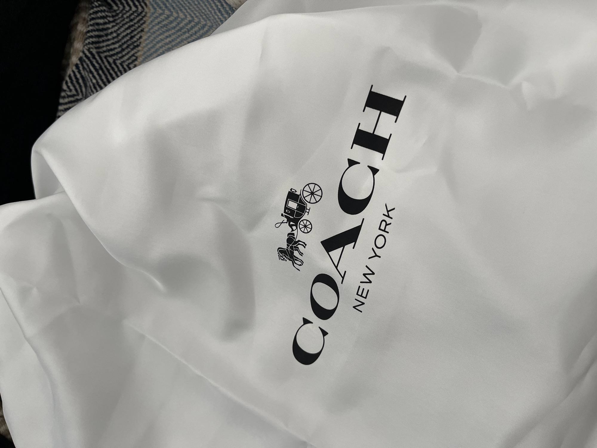 The image shows a white fabric dust bag with the black Coach logo and "NEW YORK" text printed on it. The bag is slightly creased and draped. In the background, a dark and textured material, possibly a blanket or throw with a chevron pattern, is visible. The scene appears to be indoors. The image does not contain any information about the city of Limassol, Cyprus.