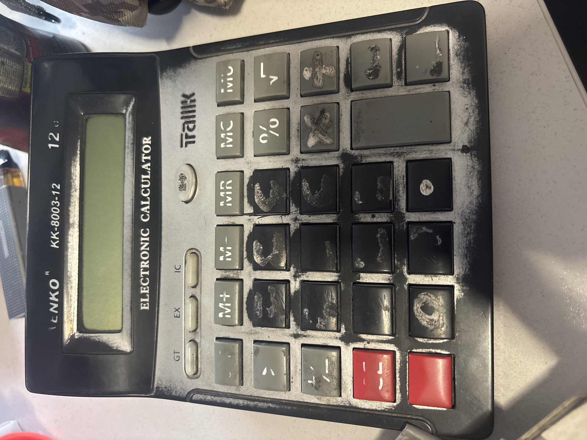 The image is a close-up, overhead shot of a black electronic calculator with a silver keypad. The calculator is slightly worn and dusty, with some of the key labels smudged. The calculator is identified as a "LENKO KK-8003-12 12C ELECTRONIC CALCULATOR". The keypad features various buttons, including numerical digits, arithmetic operations (+, -, *, /, %, sqrt), memory functions (M+, M-, MR, MC), and others like GT, EX, and IC. Two prominent red buttons are located on the left side of the keypad. The calculator is positioned on a light-colored, textured surface. The background is out of focus but appears to show other objects, including what might be a bottle and some fabric. There is no indication of the city of Kryvyi Rih, Ukraine, or any specific location within it.