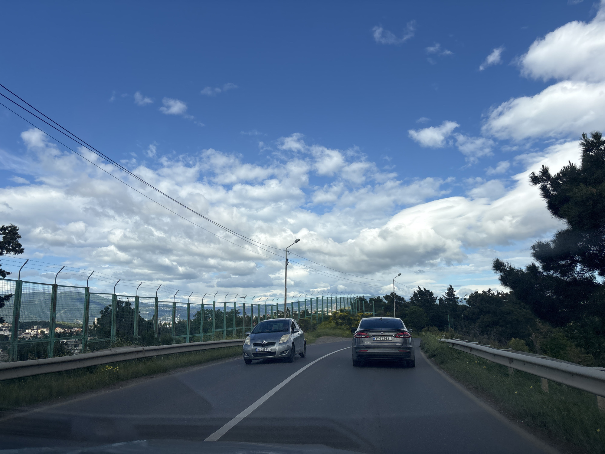 The image captures a view from inside a moving vehicle on a road in Tbilisi, Georgia. In the foreground, the asphalt road is marked with a single white line dividing the lanes. Two vehicles are visible: a silver subcompact car with license plate "HD-368-PE" in the oncoming lane, and a dark gray sedan with license plate "VV-783-EE" in the same lane as the camera. The background includes a high green fence topped with barbed wire, suggesting a protected area or border. Behind the fence, a cityscape of Tbilisi can be seen, with buildings and trees interspersed. The sky is mostly blue with scattered white clouds. Power lines stretch across the upper part of the frame. The environment is green with trees and vegetation alongside the road. The overall impression is that the road is elevated and offers a view of the city.