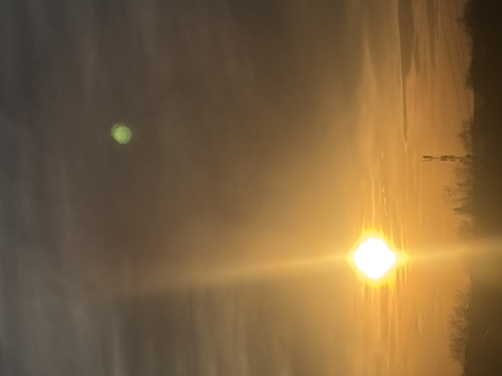 This image displays an outdoor sky scene over the city of Chaikovsky, Russia, characterized by intense solar light, suggesting either sunrise or sunset. A brilliant, white-yellow sun, presenting as a polygonal shape with a prominent vertical light flare, is centrally located in the lower half of the frame. The sky transitions from a vivid golden-orange hue near the horizon to a darker, grayish-brown overhead, with scattered, diffuse cloud formations. In the upper right quadrant, a distinct, spherical, green-yellow optical artifact exhibiting a grid-like internal pattern is visible. The foreground features a dark, silhouetted treeline. A tall, dark, indistinct structure, consistent with a telecommunications tower, along with several horizontal lines resembling power lines, is silhouetted against the bright sky on the right side, just above the horizon.