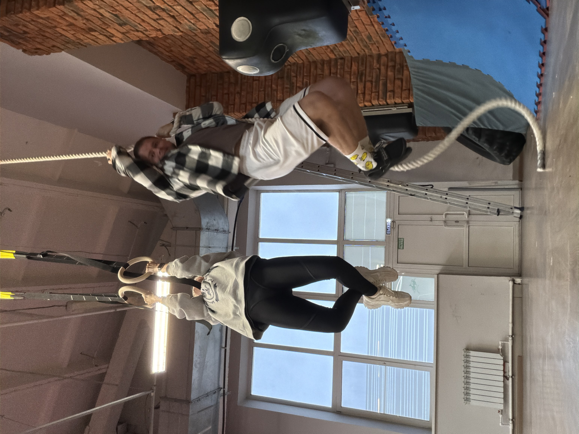 The image, taken in a gym in Chaikovsky, Russia, shows two people engaged in fitness activities. In the upper part of the image, a man with short hair, wearing a plaid shirt over a dark t-shirt, white shorts, and printed socks, is performing a rope climb. Below him, a woman in black leggings, a light-colored sweatshirt, and white sneakers is suspended from gymnastic rings. The gym environment includes brick and blue foam wall coverings, a black punching bag, a metal ladder, windows, a radiator, and a door marked with a "ВЫХОД" (Exit) sign.