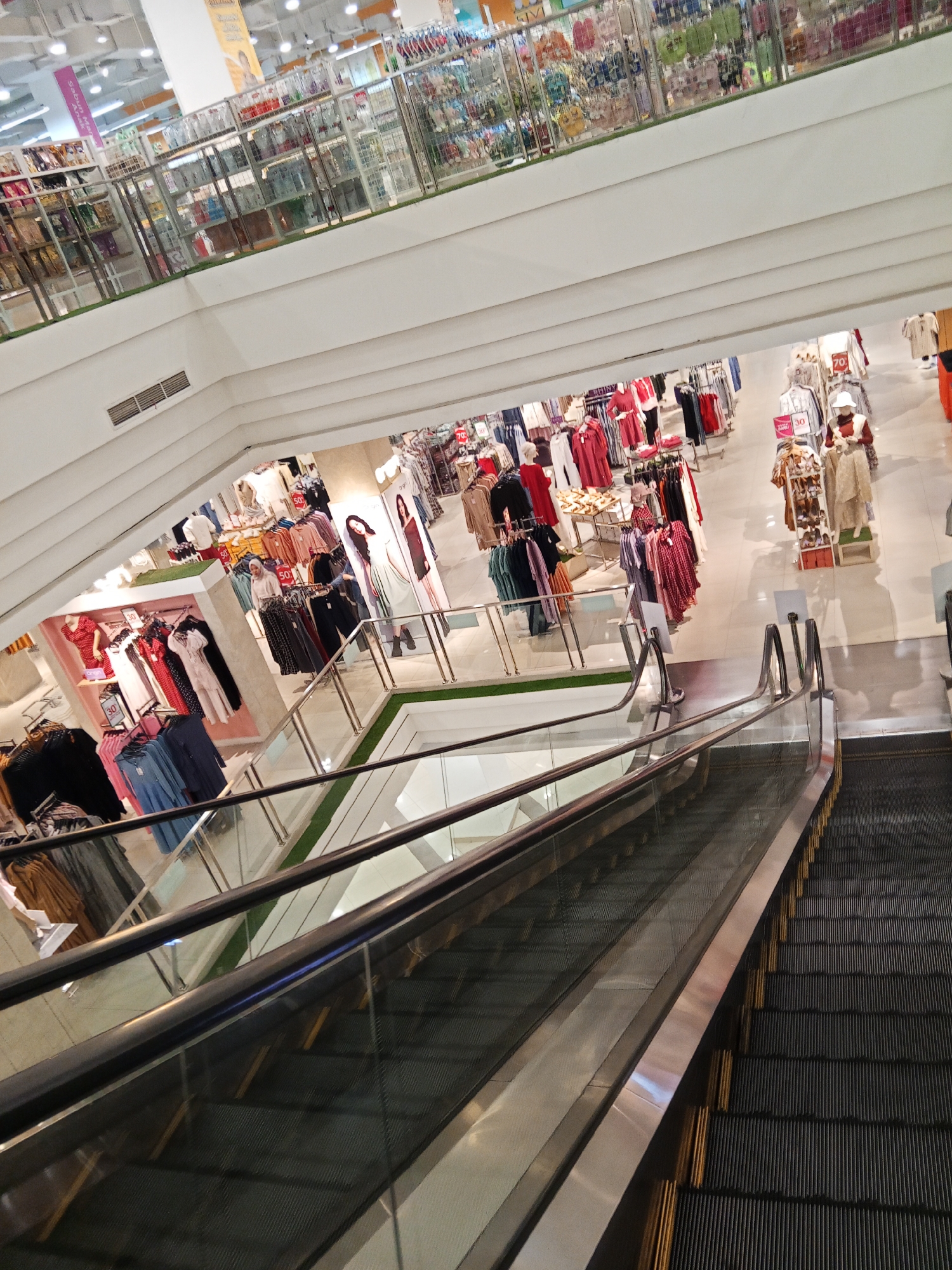 The image depicts the interior of a multi-level department store, likely a branch of Matahari Department Store in Lubuklinggau, Indonesia, given the context. The perspective is from an escalator, looking downwards into the store. The escalator's dark, ribbed steps and chrome handrails dominate the foreground. Below, a brightly lit store layout is visible, featuring clothing displays on racks and shelves. Mannequins are present, dressed in various garments. Some clothing items bear discount signs, indicating a sale. The store appears well-stocked with apparel for women, with racks of dresses, shirts, and skirts in a variety of colors. The upper levels of the store are also partially visible, showcasing more merchandise on shelves behind glass barriers. The overall impression is of a typical busy retail environment.