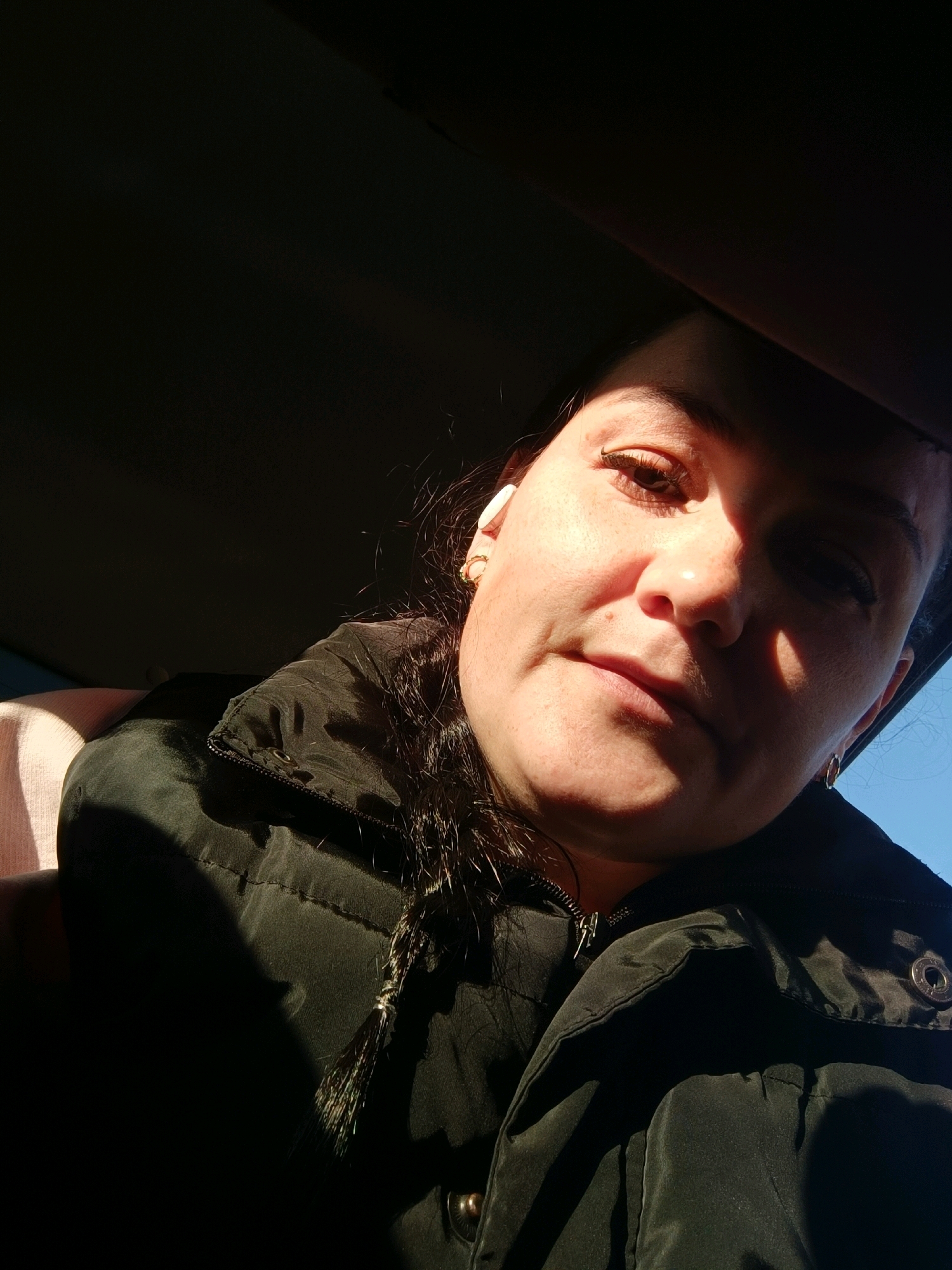 The image is a close-up, low-angle selfie of a woman with dark hair, wearing a black puffer vest over a pink garment. She has on a white earbud in her left ear and gold hoop earrings. Sunlight is casting a strong shadow across the right side of her face. The background is dark, with a sliver of blue sky visible on the right. The scene appears to be inside a vehicle, indicated by the curvature of the roof above her head and the dark interior. No specific location in Perm, Russia, can be identified.