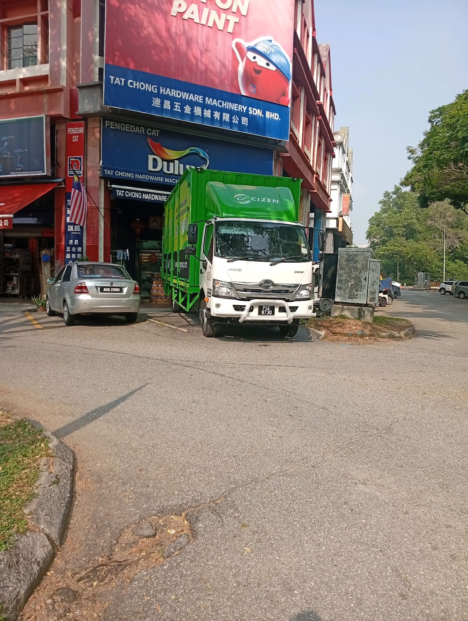 In Selayang Municipal Council, Malaysia, a green garbage truck is parked on the side of a street in front of a row of commercial buildings. The truck, labeled "Cizen," has "PSE 8730" on its front license plate. To the left of the truck, a silver sedan with license plate "AGO 2880" is parked. Above the vehicles, a red and white building displays signage for "TAT CHONG HARDWARE MACHINERY SDN. BHD." and "DULUX PAINT." The street is paved and shows some wear and tear. Trees are visible in the background. The sky is clear and blue. No people are clearly visible in the image.