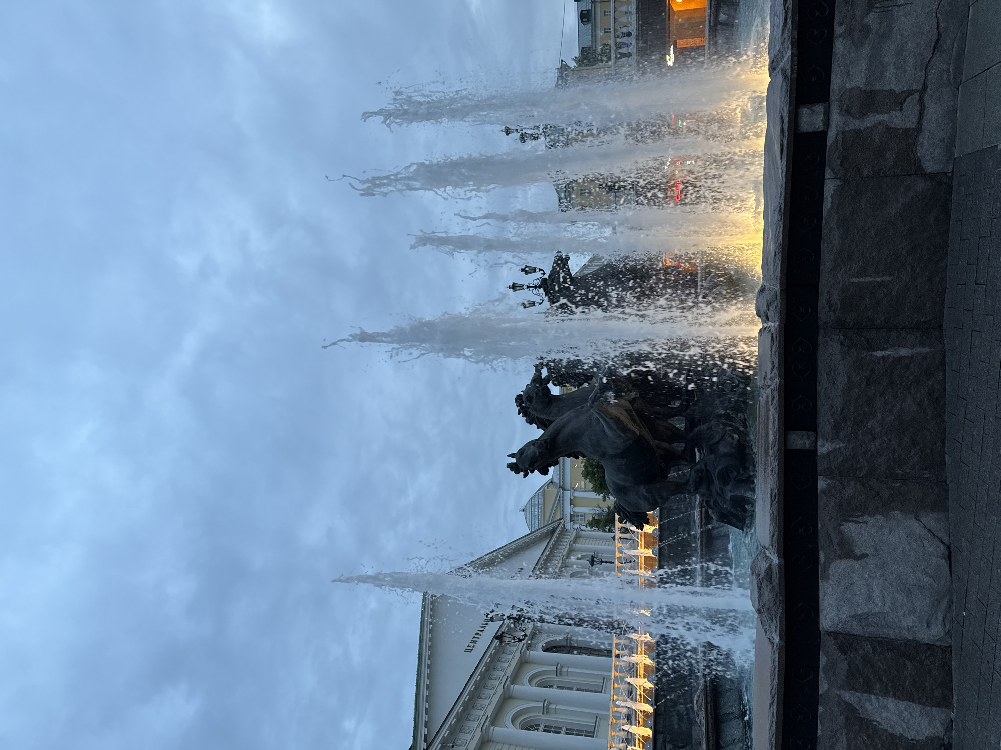 The image depicts a fountain in Moscow, Russia, featuring a sculpture of a chariot being pulled by horses. The fountain is actively spouting water. The building in the background has the word "ЦЕНТРАЛЬНЫЙ" written on it in Cyrillic. The sky is overcast with grey clouds. The frame captures a portion of the fountain's stone structure. Artificial lights are visible in the background.