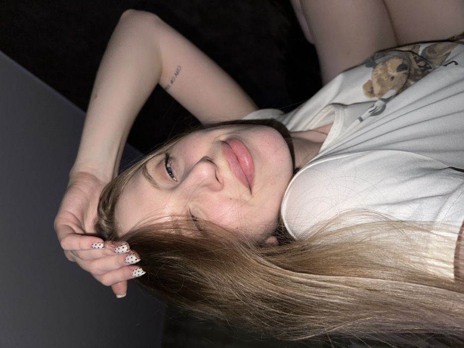 A young woman with light blonde hair and fair skin is depicted in a close-up, chest-up shot, presumably indoors in Chaikovsky, Russia. She is shown from a slight upward angle, with her head tilted, looking directly at the camera while winking with her left eye and maintaining a subtle smile. Her lips are a natural pink hue. Her right hand is raised, with fingers resting gently on the top of her head, partially touching her hair. The fingernails on her visible right hand are adorned with white polish featuring small black polka dots. She is wearing a white short-sleeved t-shirt; a brown teddy bear graphic print is partially visible on the right side of her chest. On her left forearm, a small, vertical text tattoo reads "CITY OF SUN." The background is dark and out of focus, indicating a dimly lit indoor environment without discernible details.