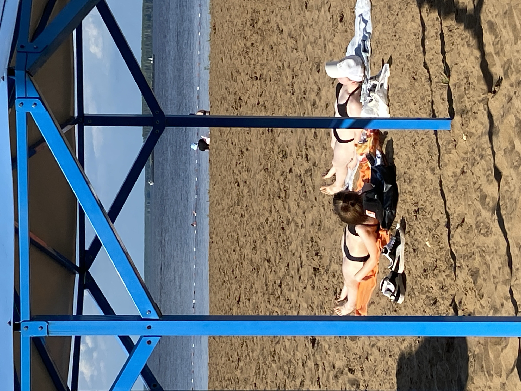 The image depicts a sunny day at a beach, likely in Chaikovsky, Russia, given the provided context. The scene is viewed from a slightly elevated perspective, looking down on two young women in swimwear on the sand. The first woman, closer to the camera, wears a black bikini and an orange sarong. She is facing away from the camera, looking towards the water. A white baseball cap adorns her head. Beside her, a second woman also wears a black bikini and a white cap, though her face is not clearly visible. Both women appear relaxed, enjoying the beach environment.
The beach itself is sandy, with the water of a lake or sea stretching out in the background. The water is calm and blue, reflecting the clear sky. The sky is partly cloudy, suggesting a pleasant, warm day. A blue metal structure, possibly a lifeguard stand or part of a beach facility, is visible in the foreground and to the left of the image, framing the scene. There are no visible text elements in the image. The overall mood is one of leisure and outdoor recreation on a summer day.