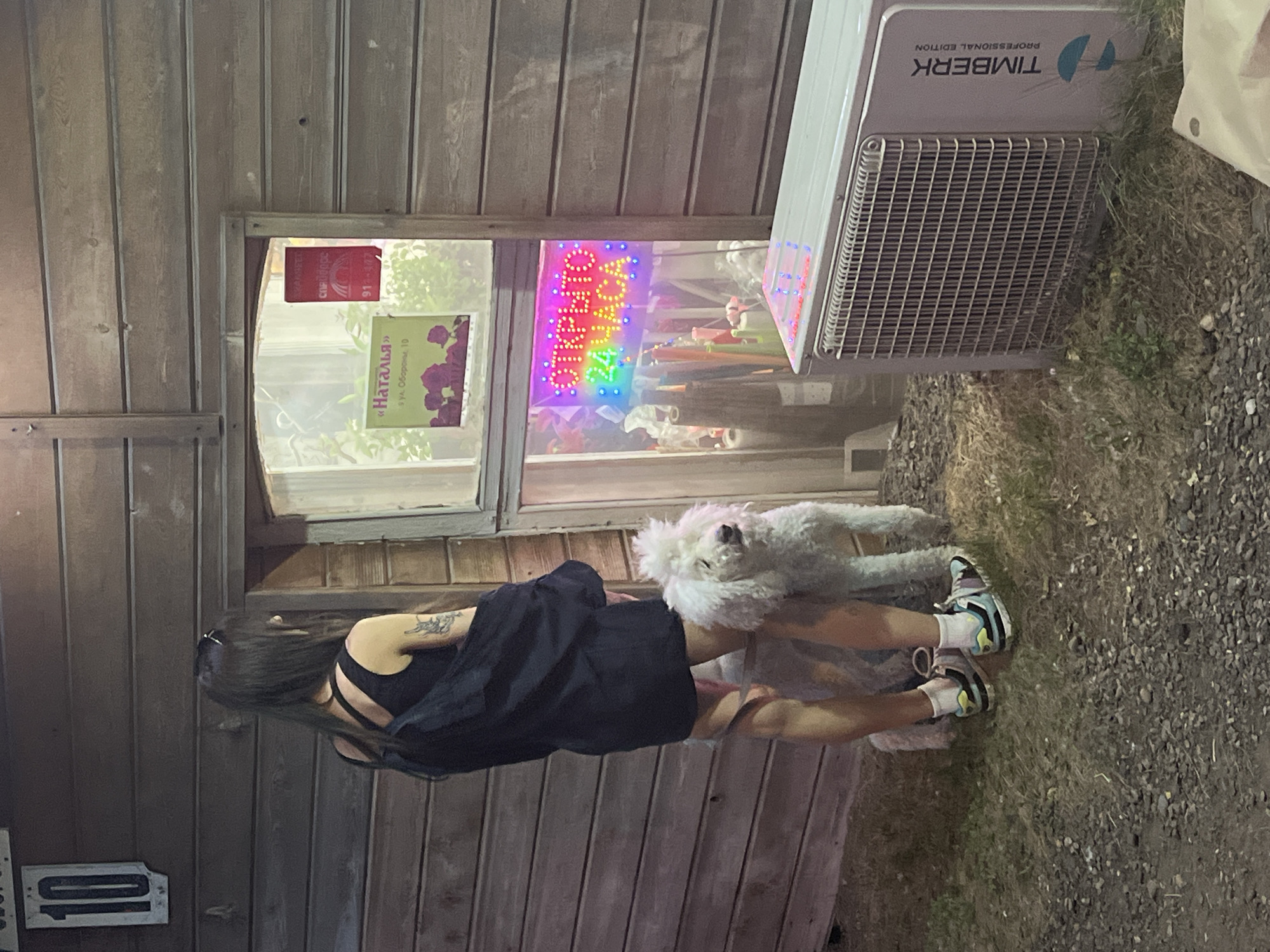 The image shows a woman and a white poodle standing outside a small wooden building. The building has a window that displays a neon sign that reads "ОТКРЫТО 24 ЧАСА" (Open 24 Hours). There is also a sign in the window that says "Наталья" (Natalia) with an address below it. The woman is wearing a black dress and sneakers, and the dog is leashed. To the right of the building, there is an air conditioning unit. The building appears to be located in a rural or semi-rural area, with gravel and grass on the ground. The overall impression is one of a small, local business.
The sign in the window provides the address: "ул. Обороны, 10" (Oborony Street, 10). This address, combined with the typical appearance of such small businesses in Russia, suggests a location in a city like Krasnoyarsk. The building itself, with its wooden construction and the style of the signage, is consistent with many commercial establishments found in smaller towns or on the outskirts of larger Russian cities.