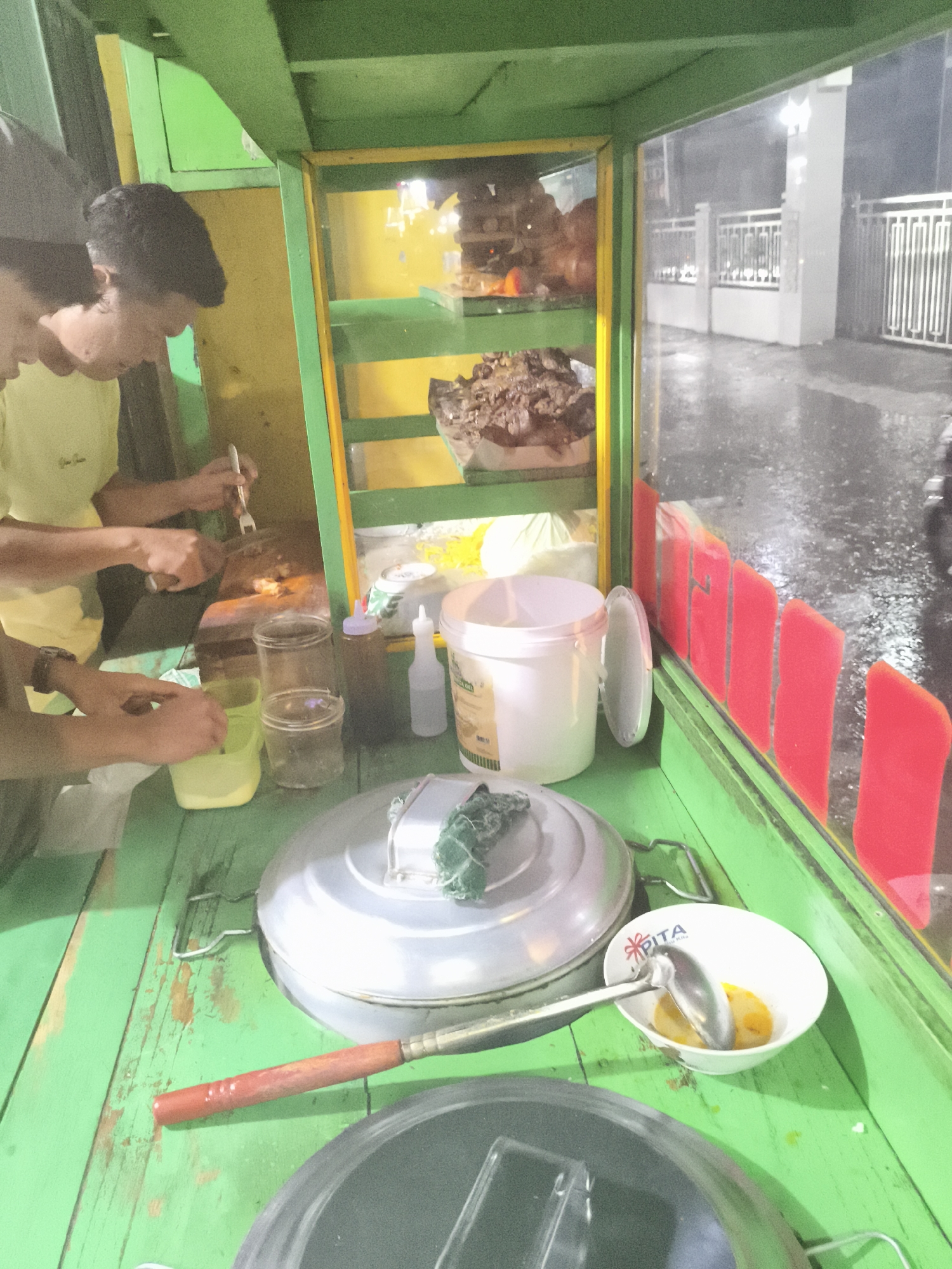 Two men are preparing food inside a green and yellow food cart, likely a street vendor setup in Depok, Indonesia. One man uses a fork to manipulate food on a dark surface, while the other handles a small container. Various containers, bottles, and cooking utensils are visible on the green countertop. Through the cart's window, a rainy street scene is visible, with a white gate and a wet road reflecting streetlights. Red decorative elements are present on the cart's side. The "PITA" logo is visible on a bowl.
