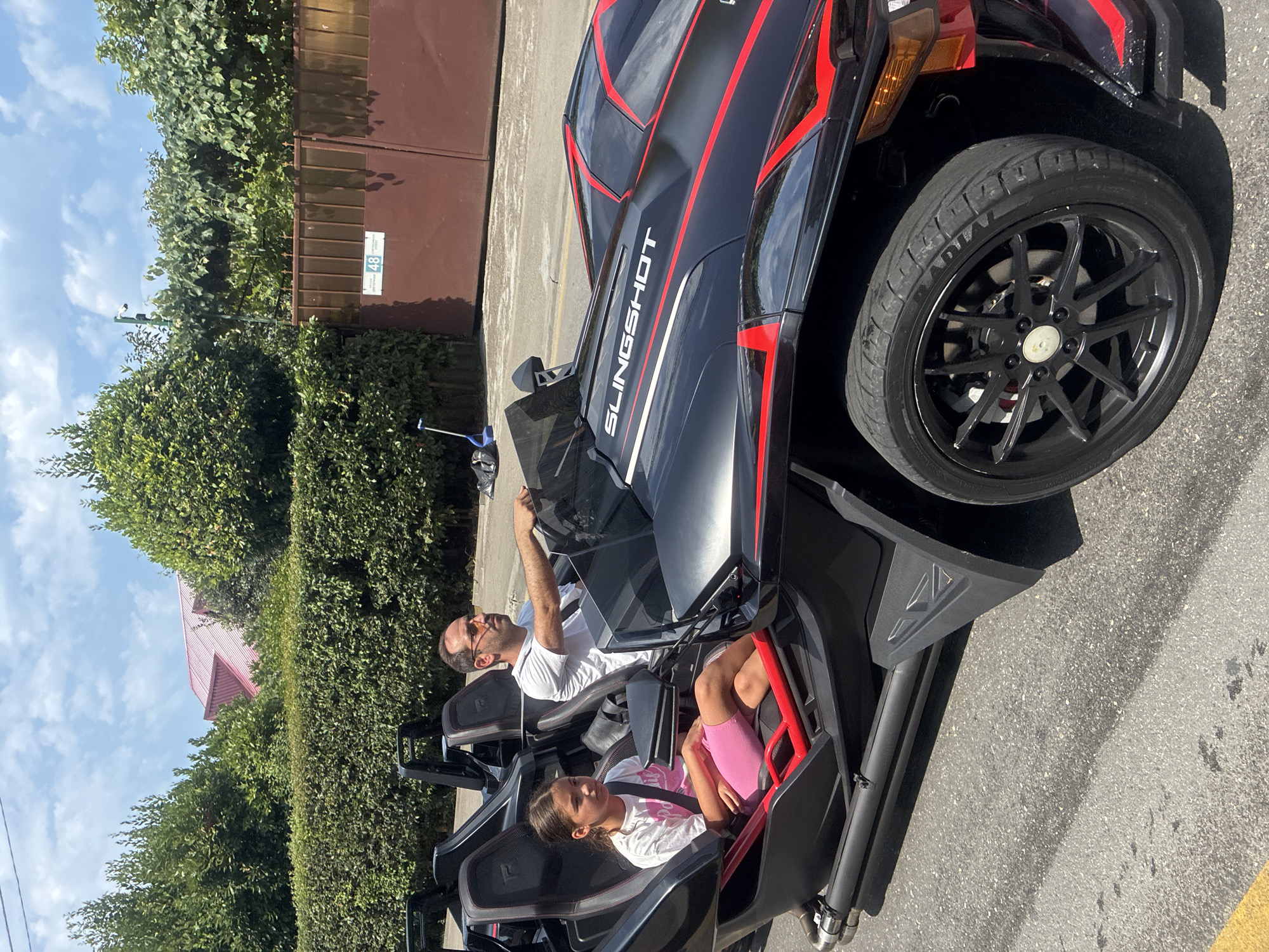 A father and daughter are riding in a black Polaris Slingshot three-wheeled vehicle on a sunny day. The father, wearing glasses and a white t-shirt, is driving. The daughter, wearing a pink top and shorts, is in the passenger seat. They are driving on a paved road with a hedge and a fence in the background. A red-roofed building is visible behind the trees. The scene is likely in Sirius, Russia, given the context.