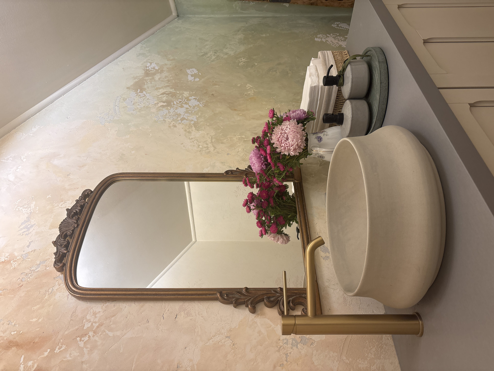 The image depicts a close-up view of a bathroom vanity area. A large, ornate gold-framed mirror hangs above a beige, bowl-shaped sink with a modern gold faucet. To the right of the sink, a bouquet of pink and purple flowers in a white pitcher with blue floral accents sits on the countertop. Behind the flowers, a stack of neatly folded white towels is visible. Two grey soap dispensers are placed on a green oval tray to the right of the towels. The countertop appears to be a dark grey material, and it sits atop a light-colored vanity cabinet with subtle detailing on the doors. The wall behind the vanity features a textured, distressed finish with a blend of beige, pink, and green tones, and there are faint floral stencils visible on the upper portion. The ceiling is angled, meeting the wall. The scene is static and well-lit, suggesting an interior shot. There are no people or animals present. The image was taken in Riga, Latvia.
