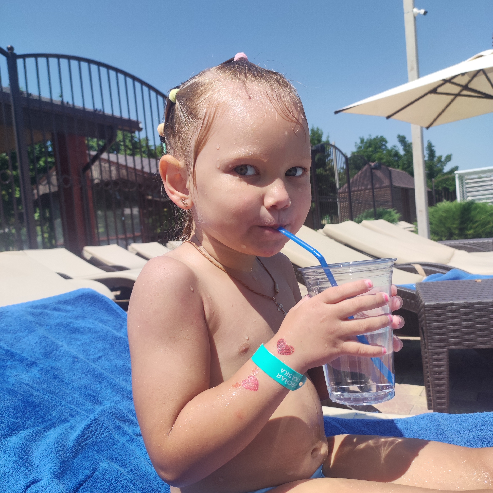 A young girl with wet hair, styled in small ponytails with colorful ties, sits on a blue towel by a pool. She is drinking from a clear plastic cup with a blue straw. She is wearing a turquoise wristband with what appears to be pink glitter hearts. There are also pink glitter hearts on her arm. She is wearing a thin necklace. The background shows lounge chairs, a fence, trees, and a white umbrella. The sun is shining brightly, indicating a warm, sunny day. The girl appears to be looking at the camera. The wristband has text in Cyrillic script, reading "ПЕСЧАНАЯ СКАЗКА" which translates to "Sandy Fairytale".