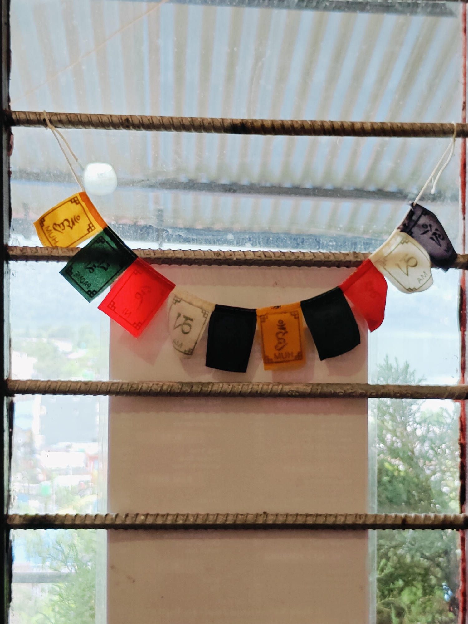 The image shows a string of prayer flags hanging across a window with metal bars. The prayer flags are colorful, with some featuring Tibetan script. The background outside the window shows a blurred view of trees and buildings, suggesting an outdoor urban or semi-urban environment. The metal bars are rusty and appear to be part of a window frame or security grate. The scene is captured from a slightly angled perspective, looking through the window. The overall impression is of a cultural element (prayer flags) placed within a functional or possibly utilitarian setting (window with bars).