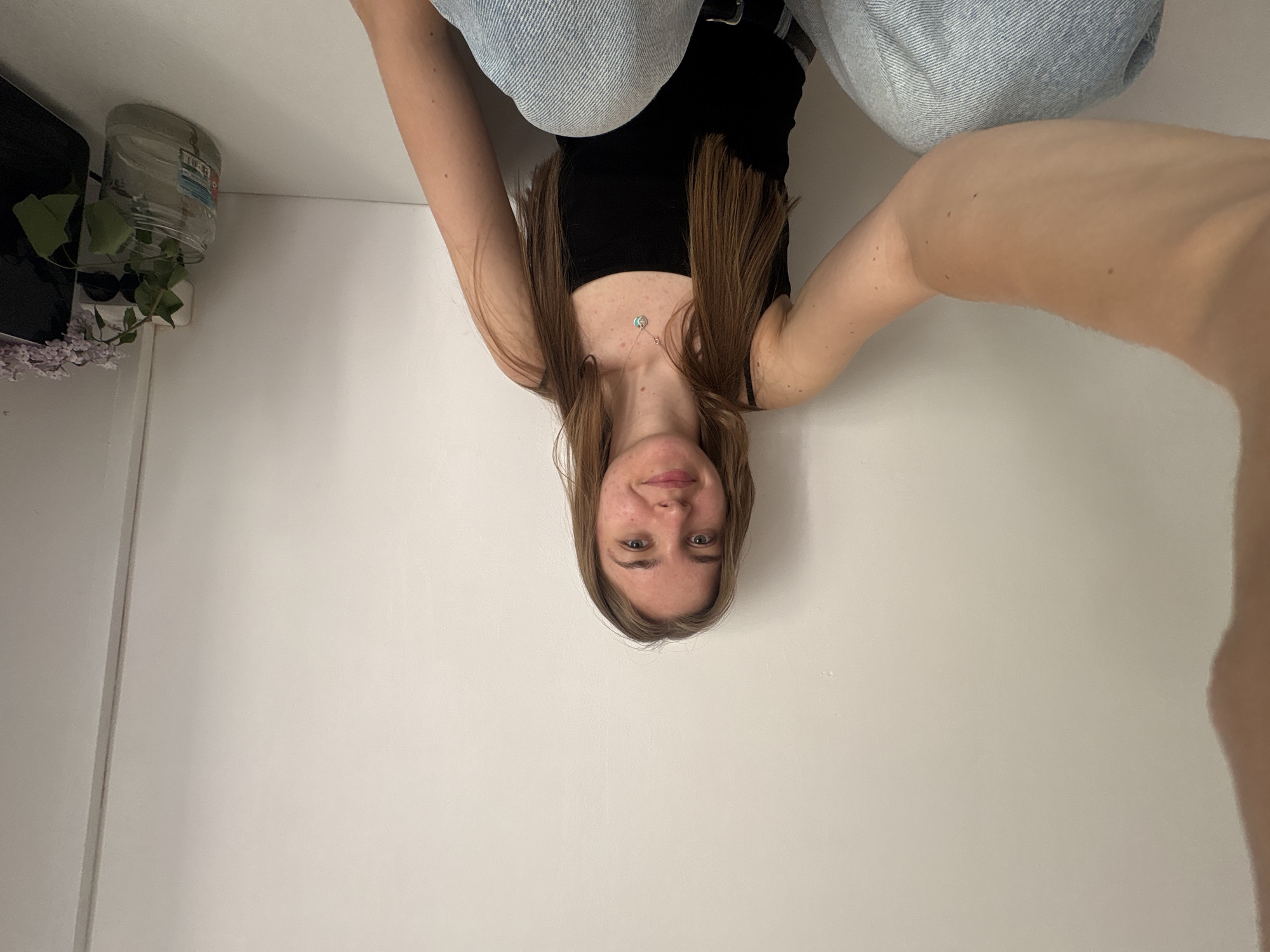 A young woman with light brown hair, wearing a black tank top and light blue jeans, is positioned upside down. Her arms are raised, presumably holding a device to take the picture. She has a necklace. The background is a white wall. To the left, there is a dark-colored object with greenery, possibly a plant in a jar, and lilac flowers. The location cannot be determined from the image.