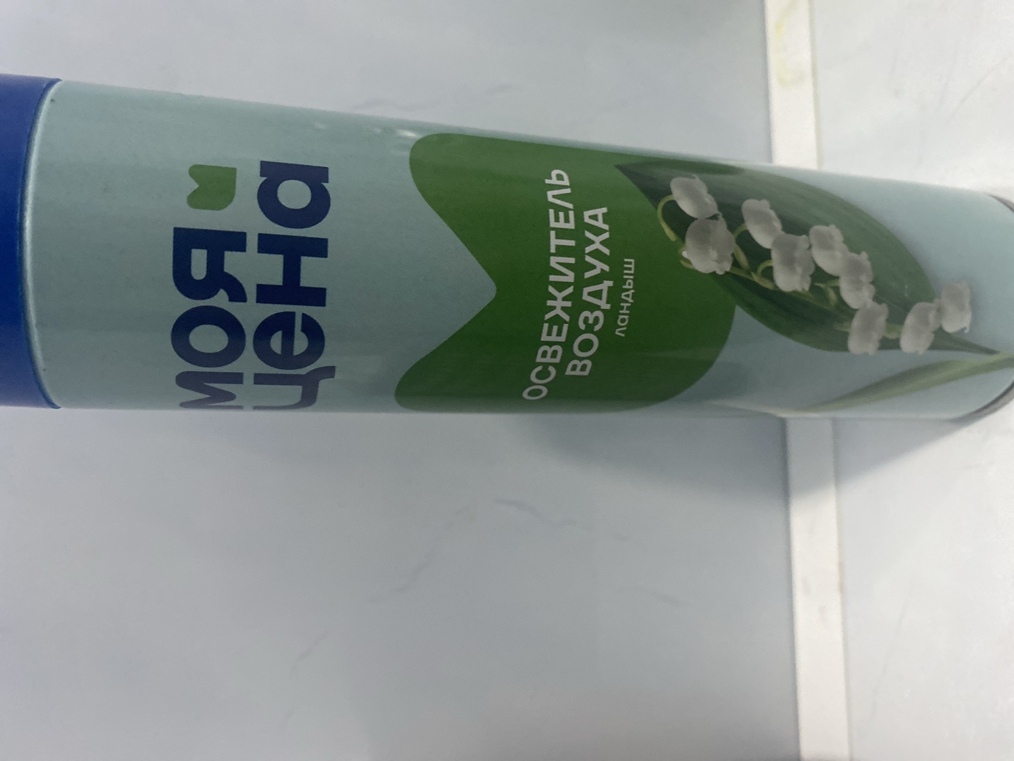 The image shows a close-up of a spray can of air freshener. The can is light blue with a darker blue cap. The label on the can features dark blue Russian text and a stylized green leaf design. The text above the leaf reads "МОЯ ЦЕНА", and below it, "ОСВЕЖИТЕЛЬ ВОЗДУХА" and "ландыш". A depiction of lily of the valley flowers with green leaves is also visible on the can. The background is a light-colored, possibly tiled wall or surface. No specific location in Kizilyurt, Russia, can be determined from the image itself.