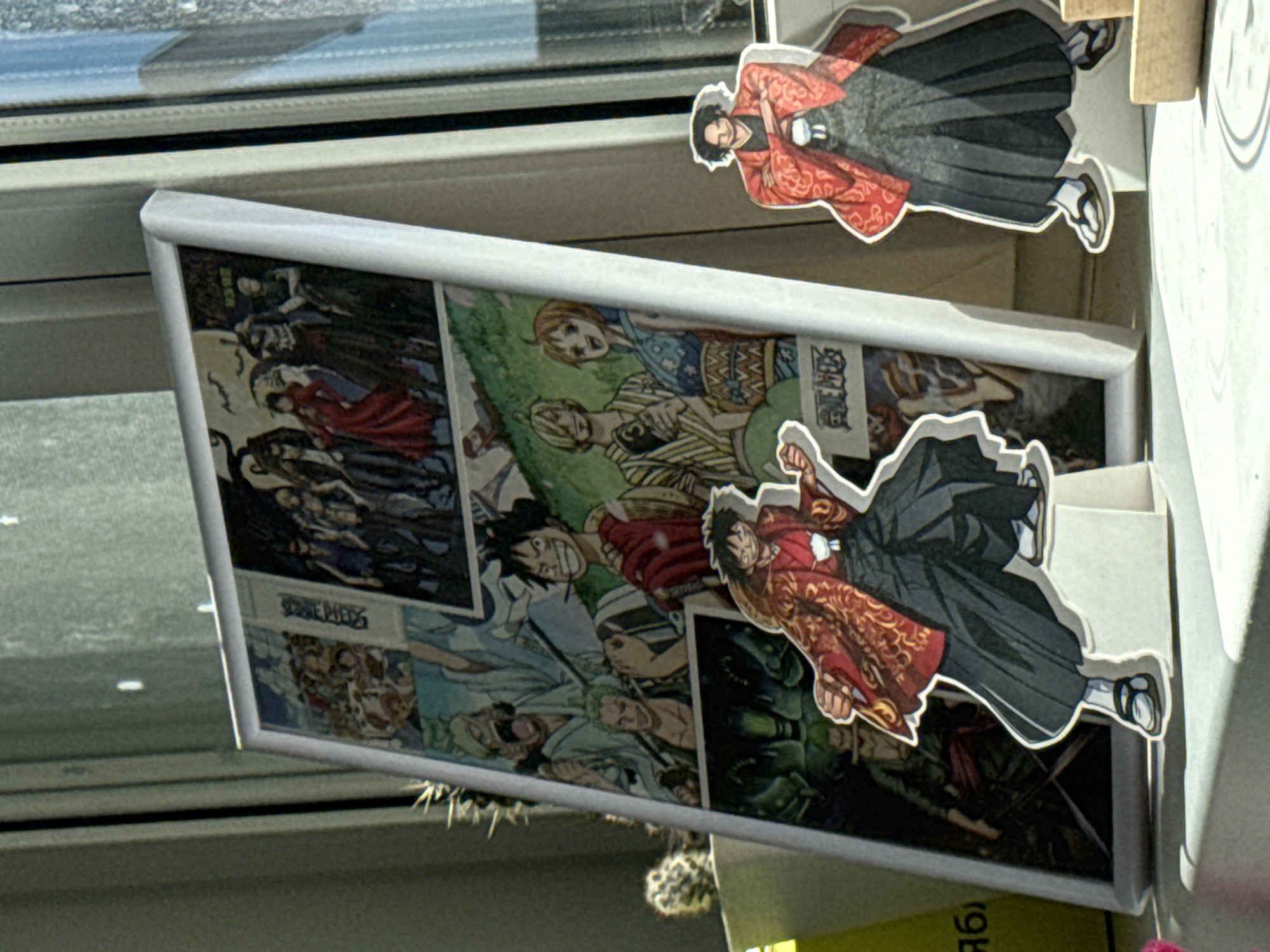 In Krasnoyarsk, Russia, a framed collage of anime characters, likely from the series "One Piece," is displayed on a windowsill. The collage features various scenes and characters from the anime in a comic-book style. To the right of the framed collage, a cutout figure of an anime character in a red kimono stands on a white stand. To the left of the collage, another cutout figure of an anime character, also in a red kimono, is positioned slightly in front. The background shows a window with a textured curtain and sunlight streaming in, creating shadows. A small potted cactus is visible to the left of the frame.