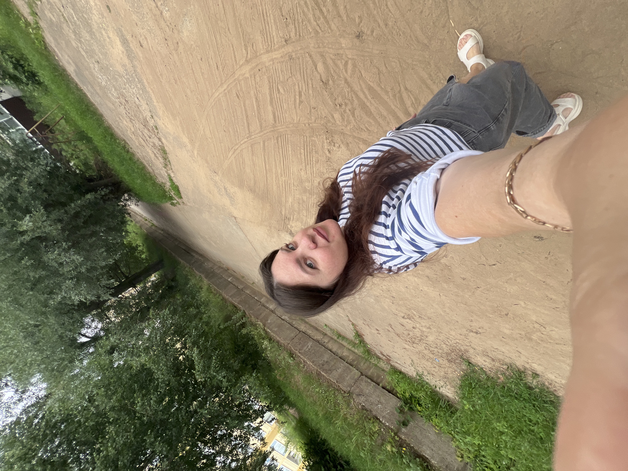 The image shows a young woman with long brown hair and blue eyes taking a selfie outdoors. She is wearing a striped t-shirt, dark pants, and white sandals. She has a bracelet on her left wrist. The woman is standing on a dirt path that slopes upwards and appears to be in an urban or semi-urban environment. To her left, there is a concrete ditch lined with grass and trees. In the background, a building with yellow siding and a playground structure are visible. The overall setting suggests a residential area. The location is identified as Noviy, Russia.